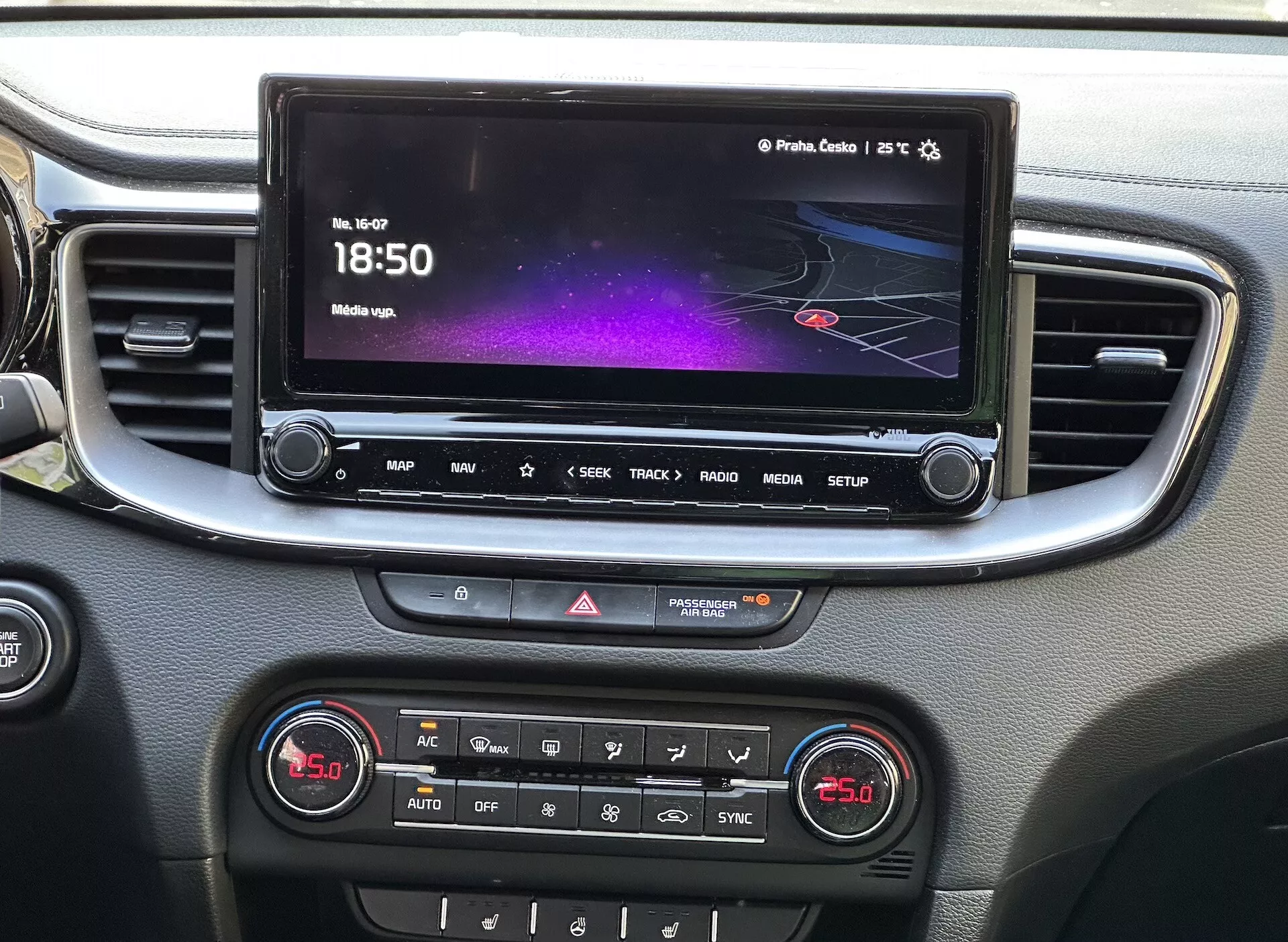Vista detallada del interior del Kia Xceed 2022 que muestra la consola central con una pantalla táctil de infoentretenimiento exhibiendo una interfaz colorida con navegación e información de medios, una fila de botones de acceso rápido, control de luces de emergencia y un panel de control climático de doble zona, todo enmarcado con un ribete plateado.