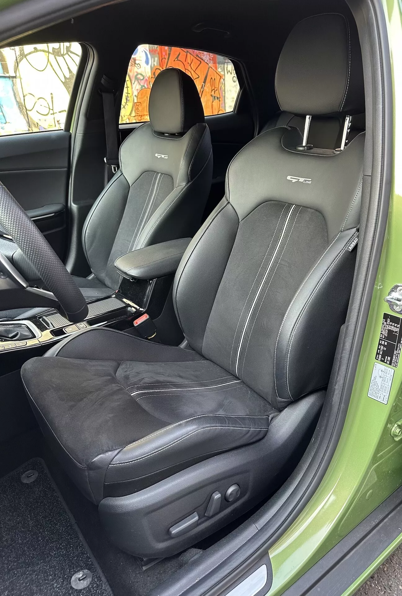 Vista interior del Kia XCeed 2022 enfocando el compartimiento del pasajero delantero tapizado en cuero con costuras intrincadas y controles de ajuste de asiento electrónicos, desde un ángulo de puerta abierta mostrando un atisbo del exterior verde vibrante.