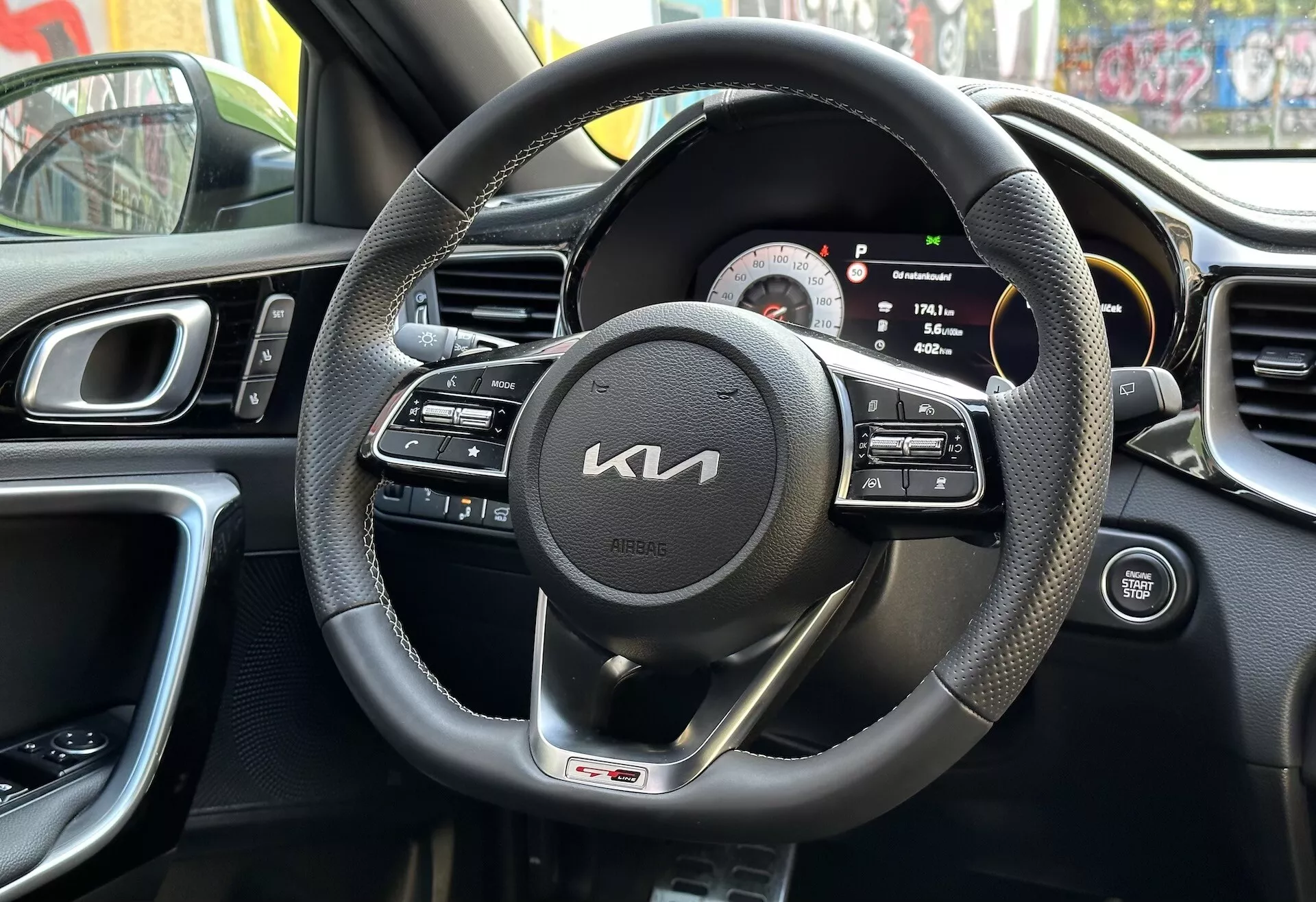 Vista interior del Kia XCeed 2022 desde la perspectiva del conductor mostrando volante envuelto en cuero negro con controles multifunción y distintivo de línea GT, clúster de instrumentos digital-analógico, parte de la consola central con ventilaciones, diseño ergonómico del tablero, y panel de la puerta del conductor con controles, reflejando entorno urbano