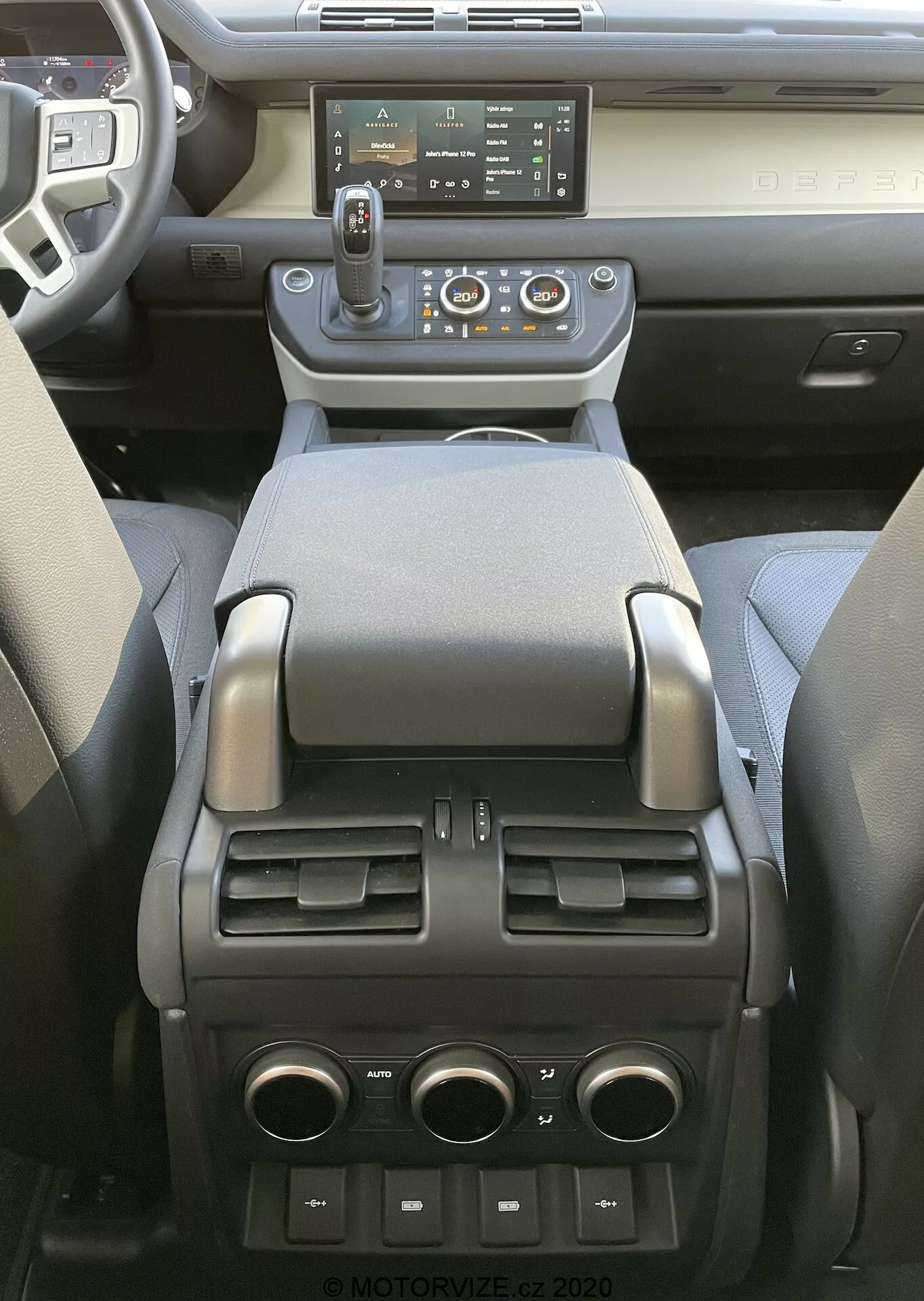 Vista interior del Land Rover Defender 2020 centrada en la consola central y la parte trasera de la cabina delantera, mostrando un sistema de infoentretenimiento de pantalla táctil moderno, ventilaciones de aire centrales con controles de climatización, palanca de cambios automática con opción de selección manual, varios botones de control, portavasos, reposabrazos suave, compartimento de almacenamiento, ventilaciones de control de clima para los pasajeros traseros, y puertos USB y un enchufe de 12V en la parte trasera, indicando una combinación de tecnología avanzada, comodidad y practicidad.