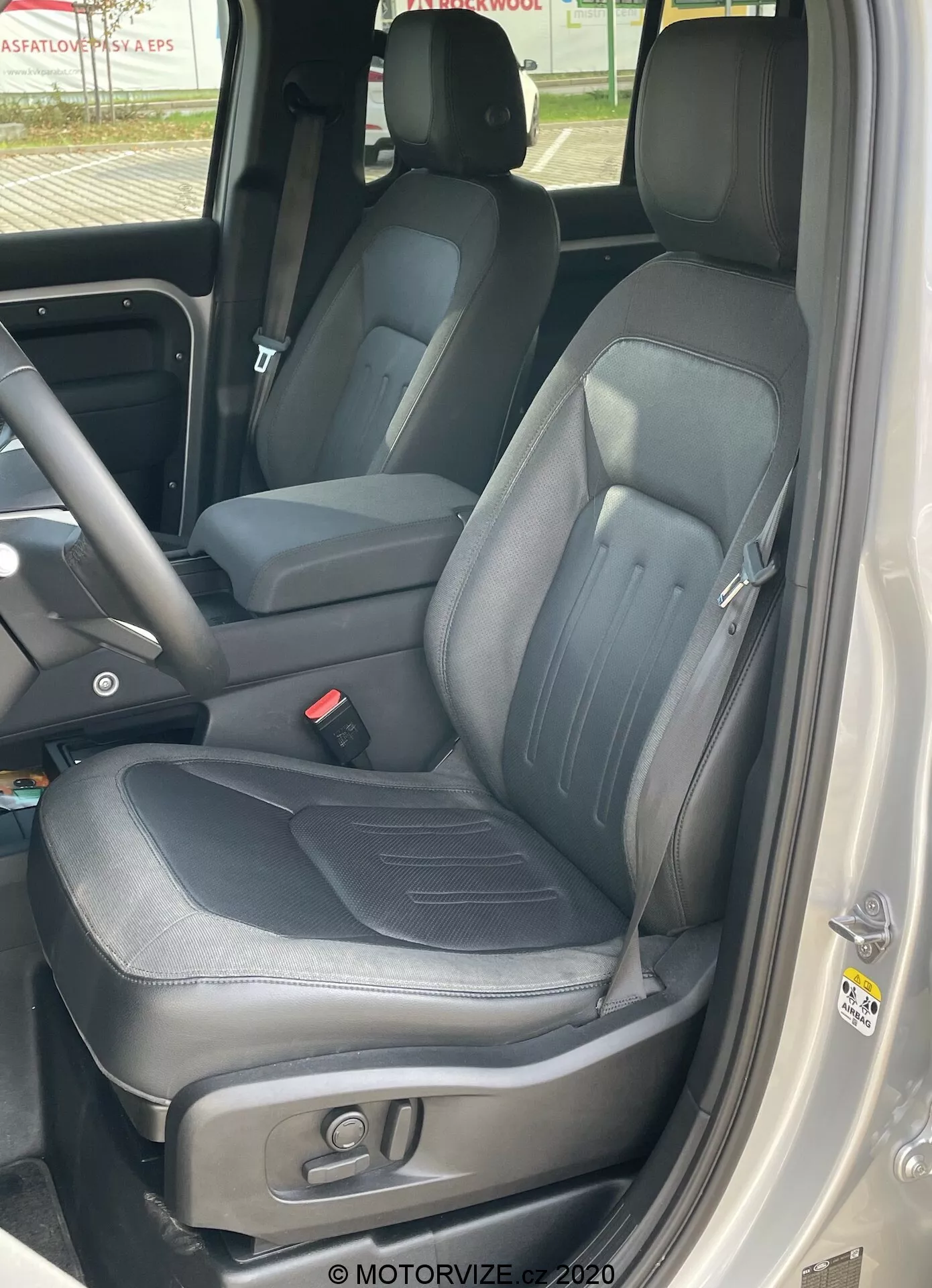 Vista de cerca del interior de un Land Rover Defender 2020 enfocándose en el asiento del conductor, tomada desde fuera del coche con la puerta abierta mostrando tapicería de cuero negro con detalles de costura visibles, refuerzos laterales, reposacabezas integrado y ajustable, controles electrónicos para ajustar el asiento, parte de la consola central que indica una cabina delantera espaciosa y el panel de la puerta con manija, botones de control y altavoces.