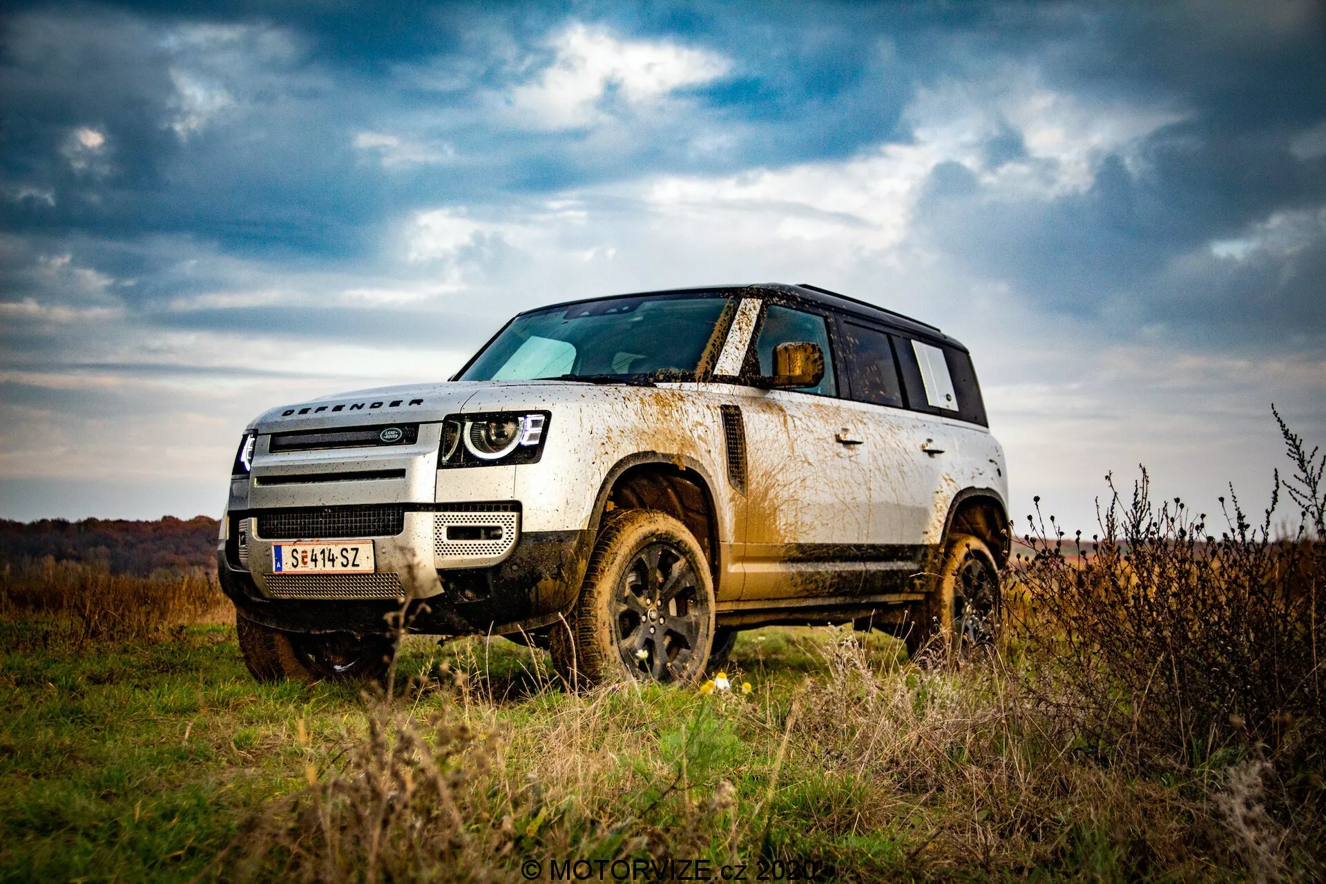 Imagen de un SUV Land Rover Defender 2020 al aire libre, destacando su capacidad todoterreno desde una perspectiva en ángulo bajo. Enfatiza la distancia al suelo del vehículo, su robustez con los arcos de las ruedas, neumáticos y elementos protectores en la parte inferior de la carrocería. Captura una vista frontal de tres cuartos, mostrando todo el frente y el lateral hasta la puerta trasera, acentuando elementos de diseño como el conjunto de faros, la parrilla y las líneas de contorno en el capó. Barro y tierra en los paneles laterales y las ruedas sugieren uso en todoterreno, indicando durabilidad y capacidad todoterreno. Cielos nublados y un paisaje otoñal en el fondo aumentan la sensación de aventura.