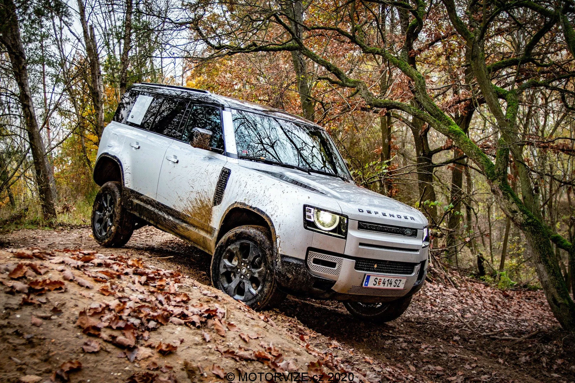 Perspectiva lateral de un Land Rover Defender 2020 SUV blanco en un entorno todoterreno; diseño distintivo con vista de la parrilla frontal y el parachoques, faros LED modernos, llantas negras de aleación con neumáticos todoterreno, espejos laterales desplegados, silueta cuadrada mostrando suciedad, manijas de puertas empotradas, línea de techo plana y alta distancia al suelo. Vehículo negociando una pendiente, mostrando sus capacidades todoterreno.