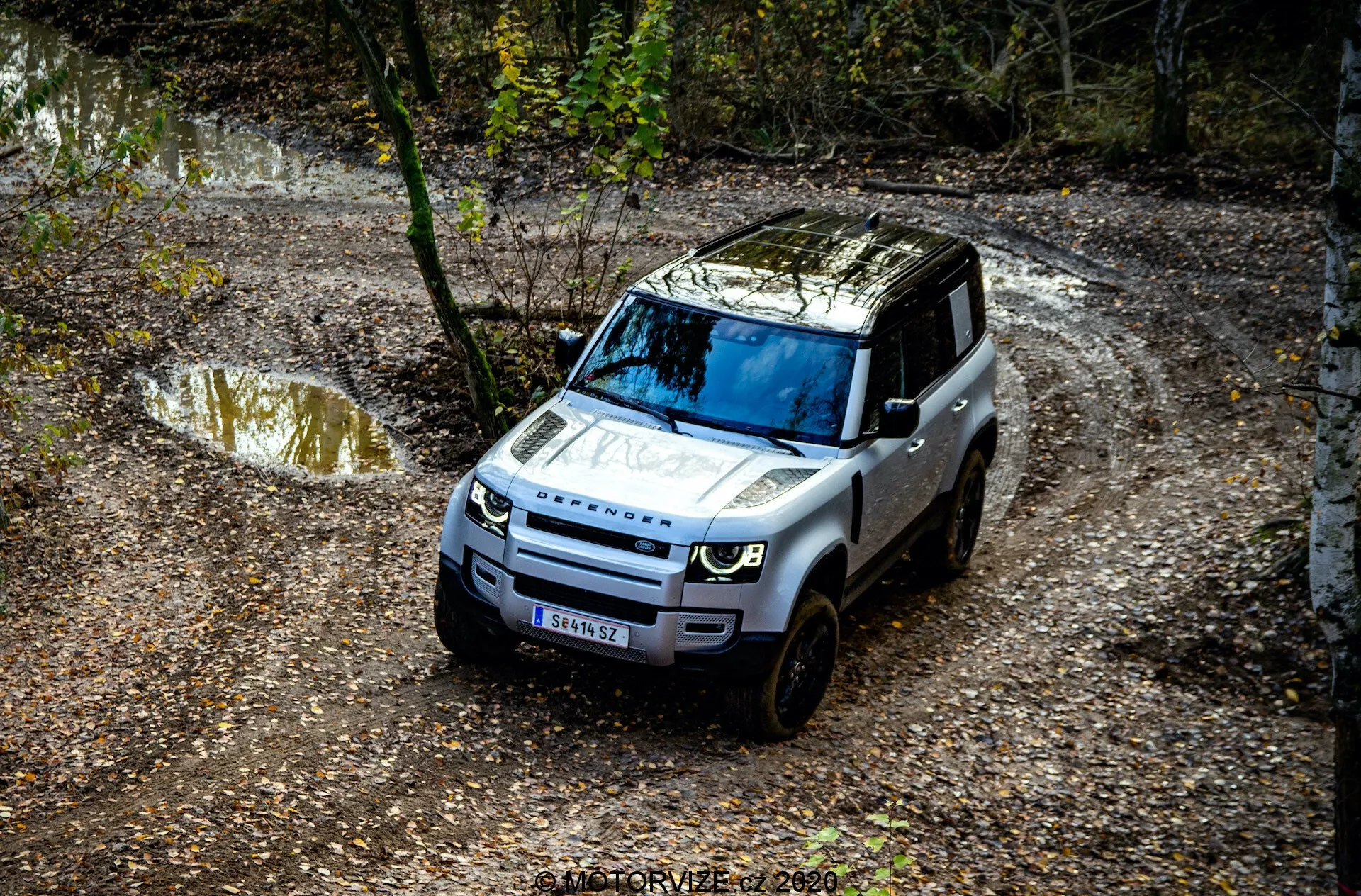 Vista de pájaro de un Land Rover Defender 2020 navegando por un sendero todoterreno, con detalles prominentes como la parrilla frontal, el capó, el parabrisas y parte del lado del conductor, rodeado por un entorno natural con charcos de agua y barro, y árboles.