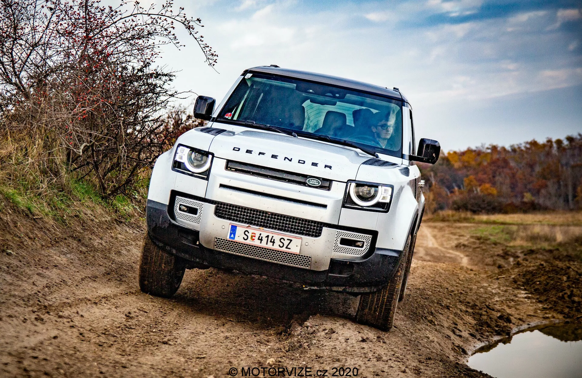 Vista de tres cuartos frontal del Land Rover Defender 2020, detalle de SUV blanco y cuadrado, todoterreno con capó plano, arcos de rueda cuadrados, luces diurnas LED circulares, faros adaptativos, parrilla de malla de panal con emblema de marca, placa protectora en el parachoques delantero, postura elevada y robusta, espejo lateral con señales de giro integradas, y énfasis en parrilla, faros y parachoques delantero. Vehículo en camino sin pavimentar con suciedad en el exterior, rodeado por entorno otoñal y terroso. Foto profesional bien iluminada con fondo borroso y marca de agua 'MOTORVIZE © 2020'.
