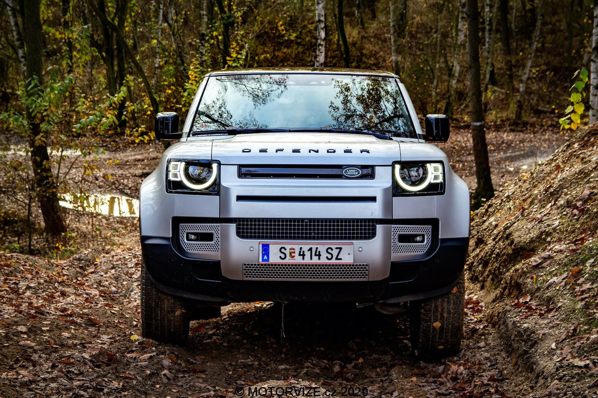 Vista frontal de un Land Rover Defender 2020 en un camino todoterreno, mostrando su presencia robusta y dominante. Los detalles visibles incluyen la parrilla distintiva con la inscripción 'DEFENDER', faros rectangulares modernos con luces diurnas LED, parachoques delantero ancho con luces antiniebla integradas, placas protectoras robustas, capó plano con bordes pronunciados, neumáticos todoterreno gruesos en ruedas de aleación y espejos laterales funcionales con intermitentes integrados. El vehículo está situado en un entorno boscoso con follaje otoñal, destacando su espíritu aventurero todoterreno.