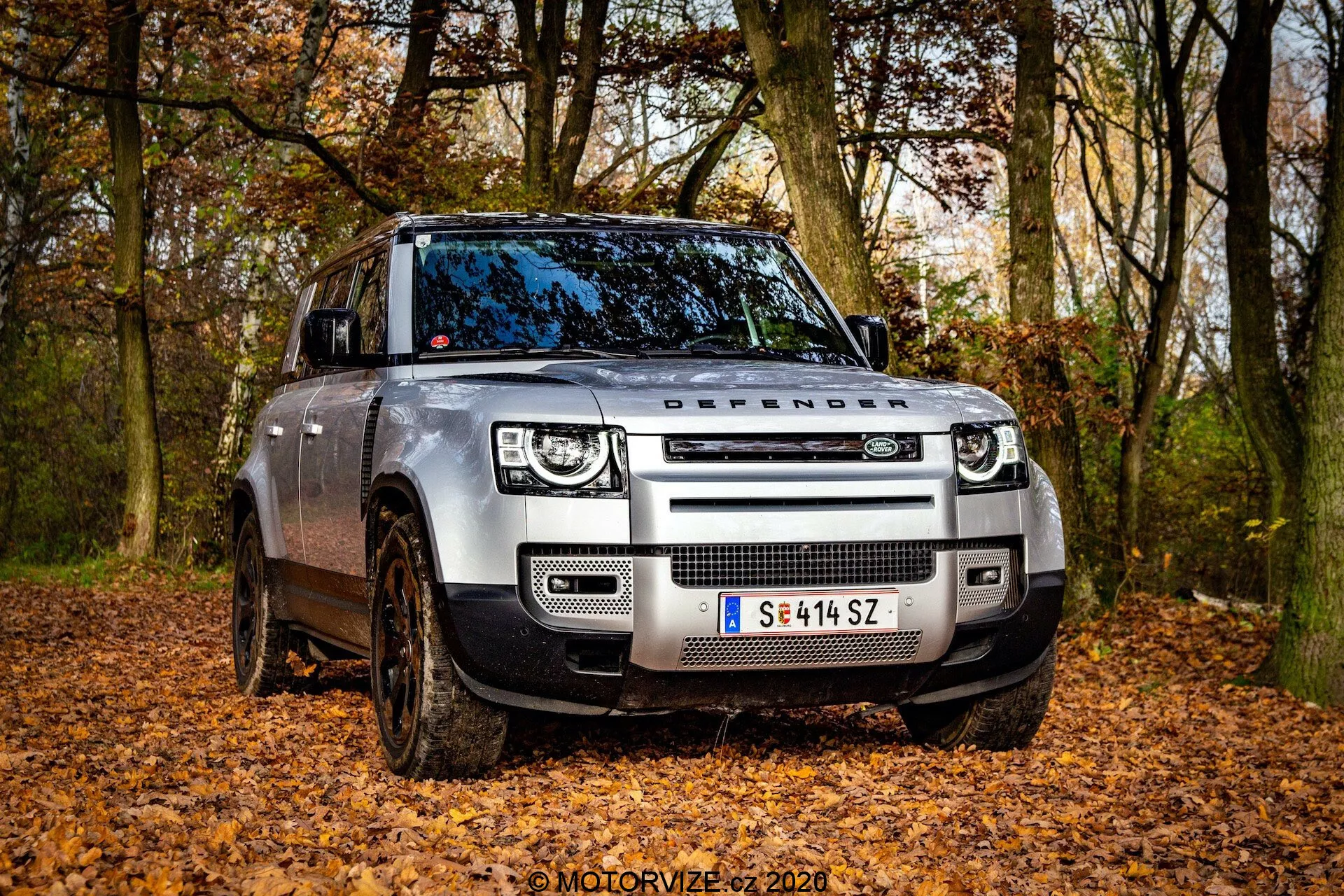 Vista de ángulo frontal de un moderno Land Rover Defender 2020 en un entorno todoterreno con hojas caídas, capturado desde una perspectiva de ángulo bajo. La imagen resalta la parrilla frontal con el texto 'DEFENDER' en el capó, los faros LED con luces diurnas cuadradas y los sensores modernos integrados en el parachoques delantero. Las ruedas cubiertas de barro y la suspensión de aire ajustable indican su capacidad todoterreno. El lado del conductor, la puerta delantera y parte de la puerta trasera son visibles, mostrando el diseño robusto del vehículo con ruedas parcialmente giradas.