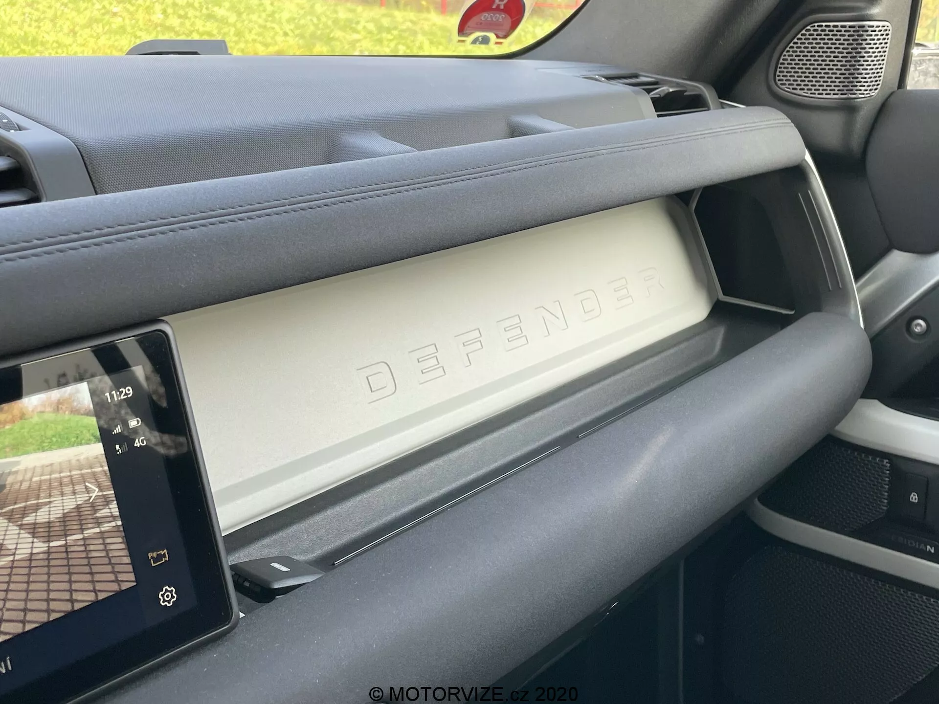 Interior del Land Rover Defender 2020 mostrando el lado del pasajero del tablero desde el ángulo del asiento del conductor. La imagen captura el compartimento de guantes cerrado con el texto en relieve 'DEFENDER', ejemplificando el diseño lujoso y resistente del vehículo. A la derecha, una pantalla táctil moderna de infotainment muestra controles del reproductor de medios y un mapa de navegación, indicando características tecnológicas avanzadas. Los materiales del tablero demuestran una combinación de texturas premium y prácticas, y la rejilla del altavoz es visible a la izquierda, integrándose con el diseño robusto del interior.