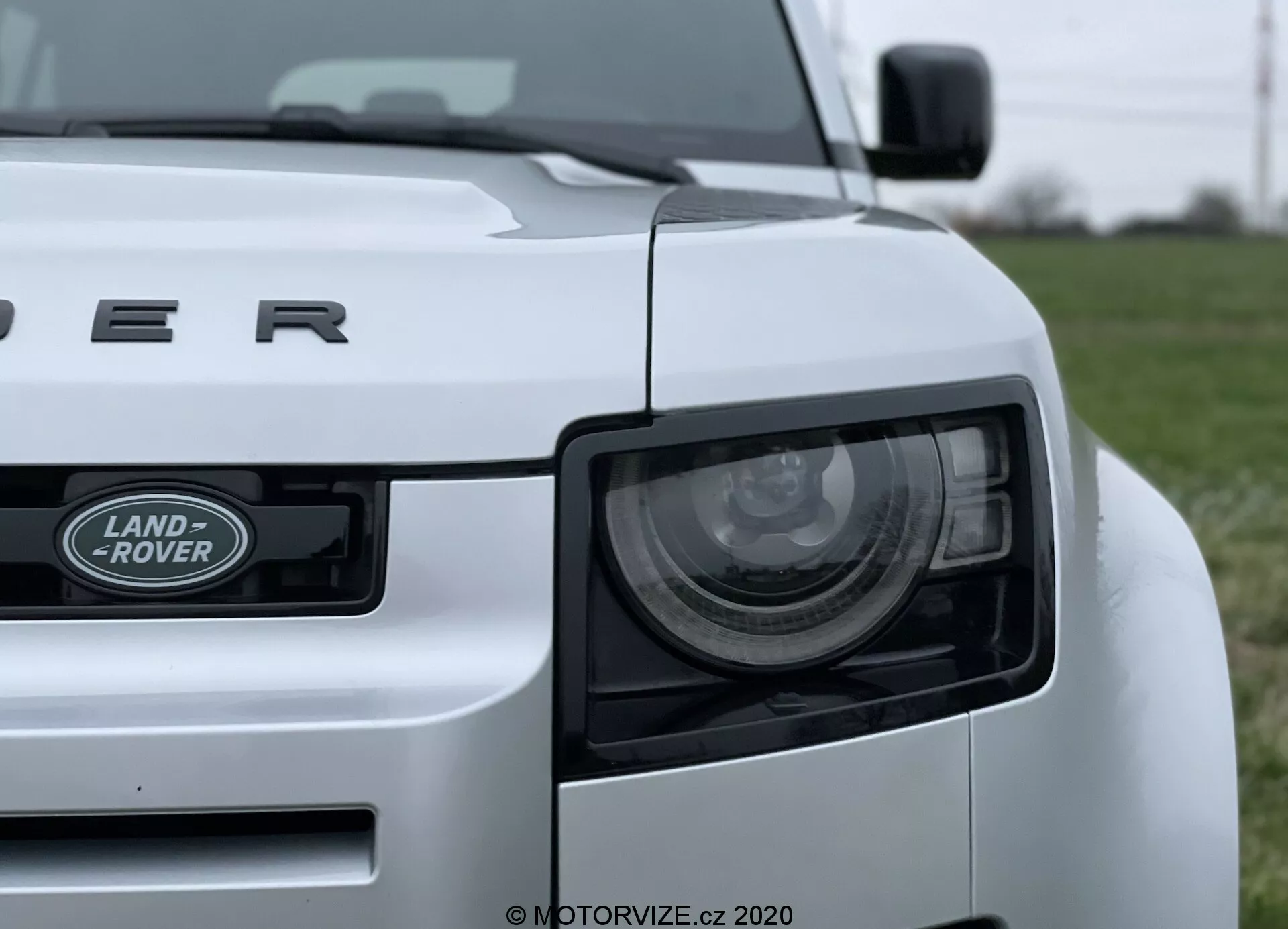 Imagen en primer plano de un coche Land Rover Defender 2020 blanco tomada desde un ángulo bajo, que muestra el cuarto delantero izquierdo del vehículo. Entre las características destacadas se incluyen el faro, la parrilla con el emblema 'LAND ROVER' y el capó con la inscripción 'DEFENDER'. El faro presenta elementos LED modernos, y el diseño robusto del coche se resalta con líneas y curvas visibles. El coche está posicionado en un campo de hierba, con el fondo desenfocado para enfatizar en el vehículo.