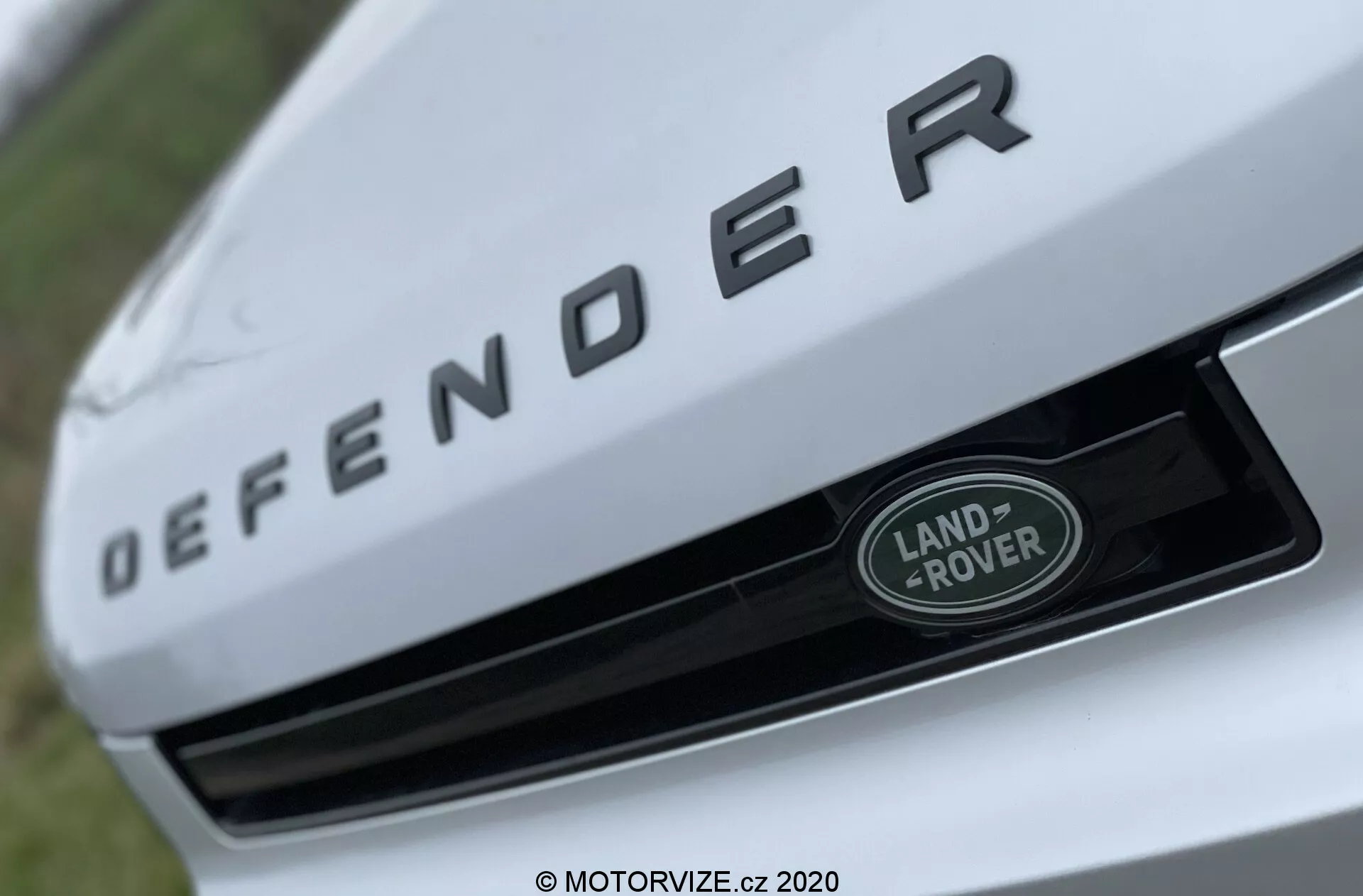 Vista de primer plano de la sección frontal de un Land Rover Defender 2020, con un enfoque destacado en la placa con el nombre 'DEFENDER' en letras plateadas en relieve en el borde frontal del capó. Debajo de la placa de identificación, la insignia de Land Rover en plata y verde con un borde blanco está posicionada centralmente en la parrilla. La imagen está tomada desde un ángulo bajo y ascendente con una profundidad de campo reducida, destacando los elementos de la marca mientras se difumina el fondo.