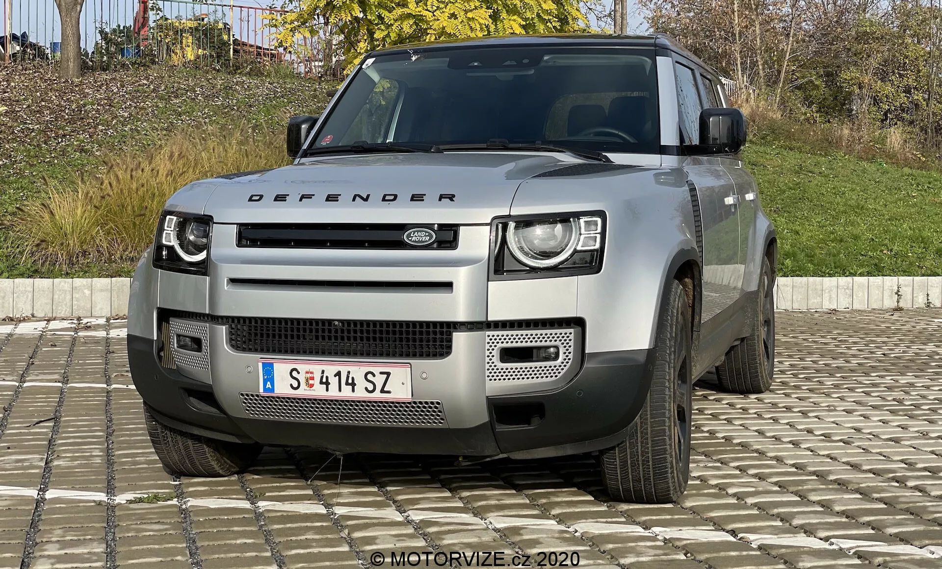 Vista frontal de un Land Rover Defender del modelo 2020, que destaca sus distintivos faros LED de dos niveles, una parrilla prominente con el logo de Land Rover, un parachoques geométrico con placa protectora plateada y otras características robustas que sugieren capacidades todoterreno. También son visibles el capó con el nombre 'Defender', las entradas de aire y vistas parciales de las ruedas de cinco radios. El vehículo es de color neutro claro, probablemente plateado o gris.
