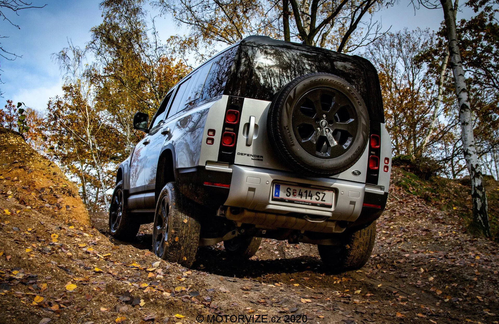 Vista trasera en tres cuartos del SUV Land Rover Defender 2020 en un terreno todoterreno, mostrando neumáticos robustos, gran altura al suelo y rueda de repuesto trasera en un entorno forestal de otoño.