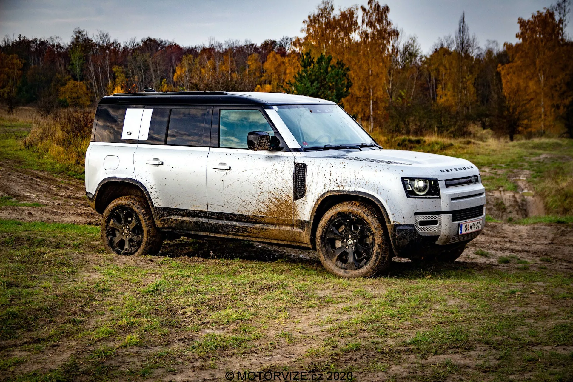 Vista lateral de un SUV todoterreno Land Rover Defender 2020 mostrado en un ángulo de tres cuartos, con su carrocería blanca con techo y ruedas negras, cubierto de barro. Vehículo posicionado en diagonal de la esquina inferior izquierda a la superior derecha contra un terreno natural embarrado bajo cielos nublados con árboles otoñales en el fondo.
