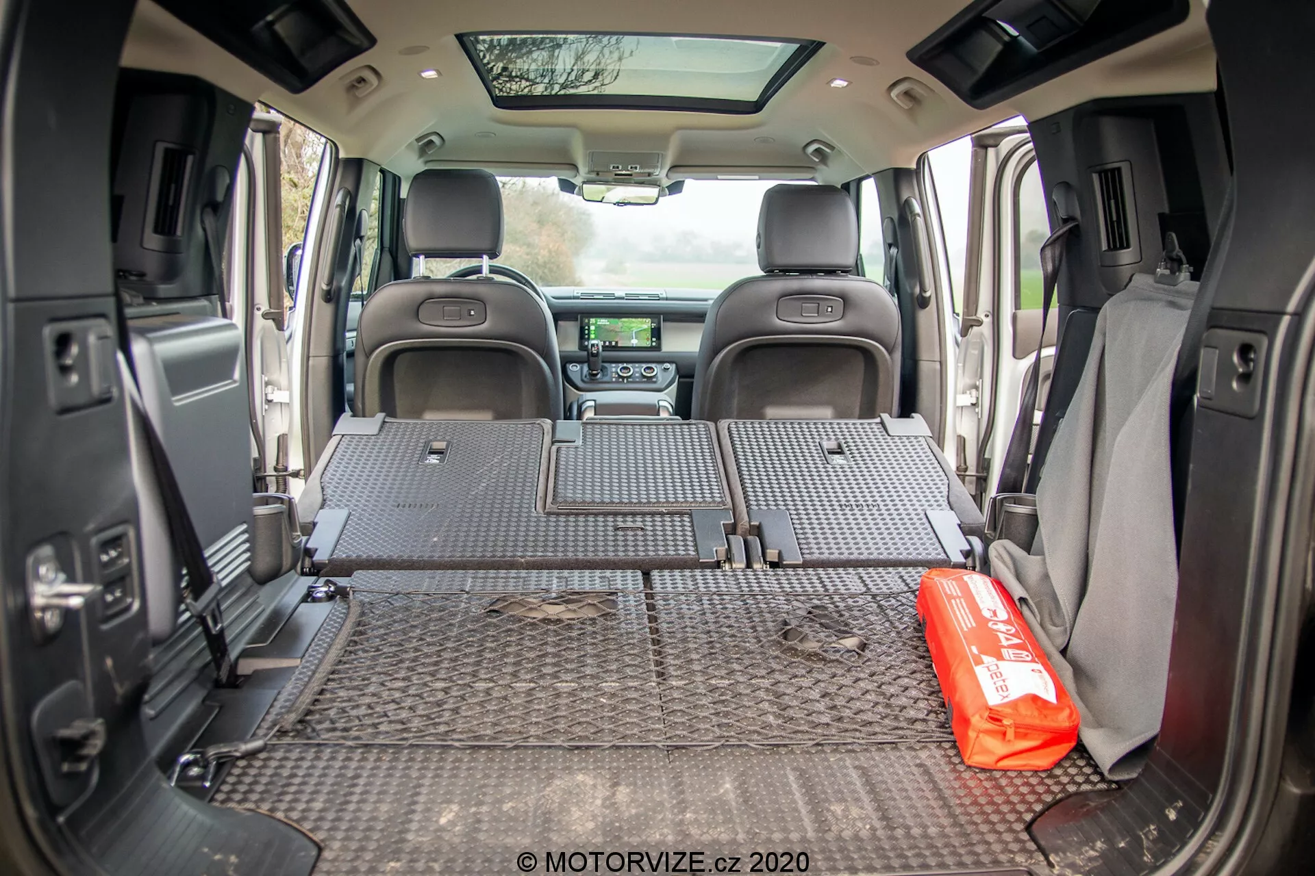 Vista interior del espacio de carga del Land Rover Defender 2020 tomada desde un ángulo orientado hacia la parte trasera, mostrando la alfombra protectora con patrones de placas de diamante que cubre todo el piso del área de carga. Los asientos traseros están plegados y planos, revelando un espacioso área de carga. Las paredes laterales tienen accesorios como altavoces y portavasos, lo que sugiere un diseño equilibrado entre funcionalidad y comodidad. El tablero frontal en la distancia es parcialmente visible, con el sistema de infoentretenimiento y controles de clima. Un kit de emergencia rojo está colocado de manera prominente en el piso del área de carga, insinuando el uso del vehículo para familias o aventuras. La puerta trasera abierta permite que la luz natural entre al área de carga.
