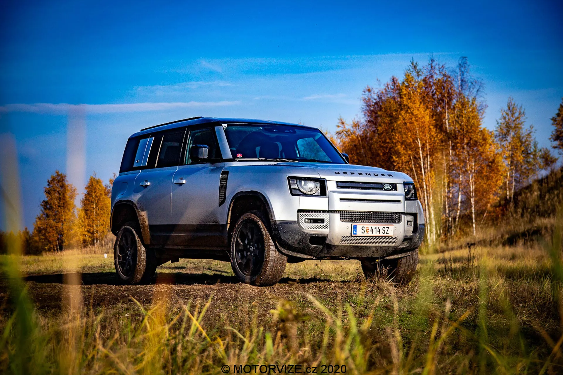 Imagen de un Land Rover Defender plata 2020 desde una perspectiva a nivel del suelo, capturada en una vista frontal oblicua en tres cuartos. El vehículo muestra prominentemente su parrilla, capó, parachoques delantero y diseño de ruedas, ligeramente orientado hacia el lado del conductor. El Defender está en un terreno exterior con un fondo de árboles otoñales con hojas rojas y naranjas bajo un cielo azul parcialmente nublado. Las características de diseño notables incluyen su distintiva forma cuadrada, faros LED modernos, luces antiniebla, parrilla de dos barras con la marca LAND ROVER, placa protectora delantera, llantas de aleación negras con neumáticos todoterreno y un techo negro contrastante con tapas de espejos negras a juego. La foto es muy nítida y dinámica, enfatizando la robustez y estética moderna del vehículo.