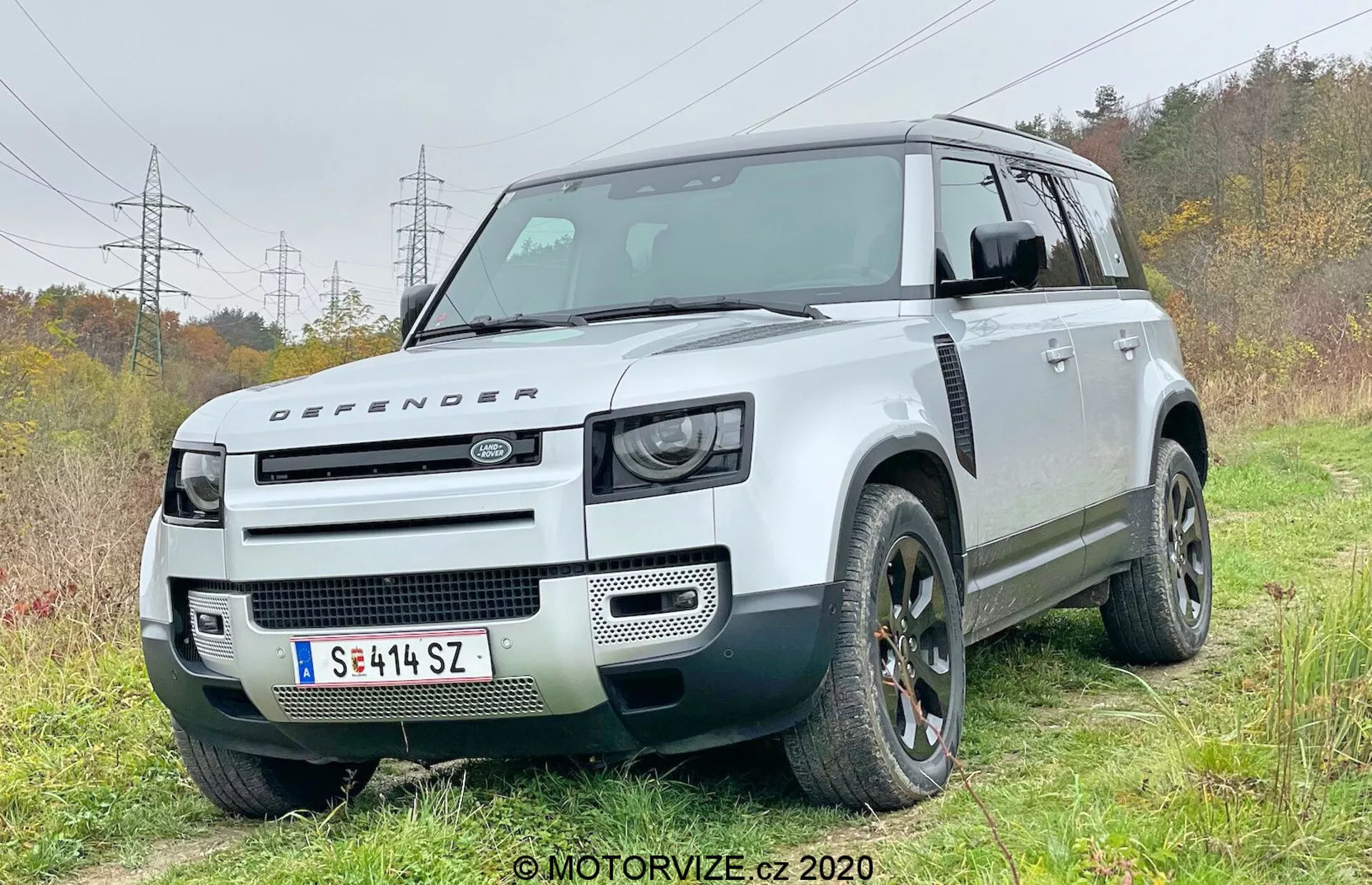 Esta fotografía captura un SUV Land Rover Defender 2020 moderno desde un ángulo de cuarto delantero, mostrando el lado del conductor. El vehículo está ligeramente fuera de carretera en una zona de césped con una pista de tierra que muestra sus capacidades todoterreno. El diseño distintivo y cuadrado tiene una alta distancia al suelo, un esquema de color en dos tonos con carrocería blanca y techo negro contrastante. La fascia frontal muestra conjuntos de faros LED de inspiración retro, un capó con la insignia Defender y una parrilla frontal minimalista, destacando su reputación todoterreno robusta. Las características técnicas visibles incluyen sensores de estacionamiento delanteros integrados en el parachoques, sugiriendo sistemas avanzados de asistencia al conductor. Los arcos de rueda robustos acomodan neumáticos grandes todoterreno, y elementos de diseño como ruedas de aleación y diseño de parrilla único se destacan.