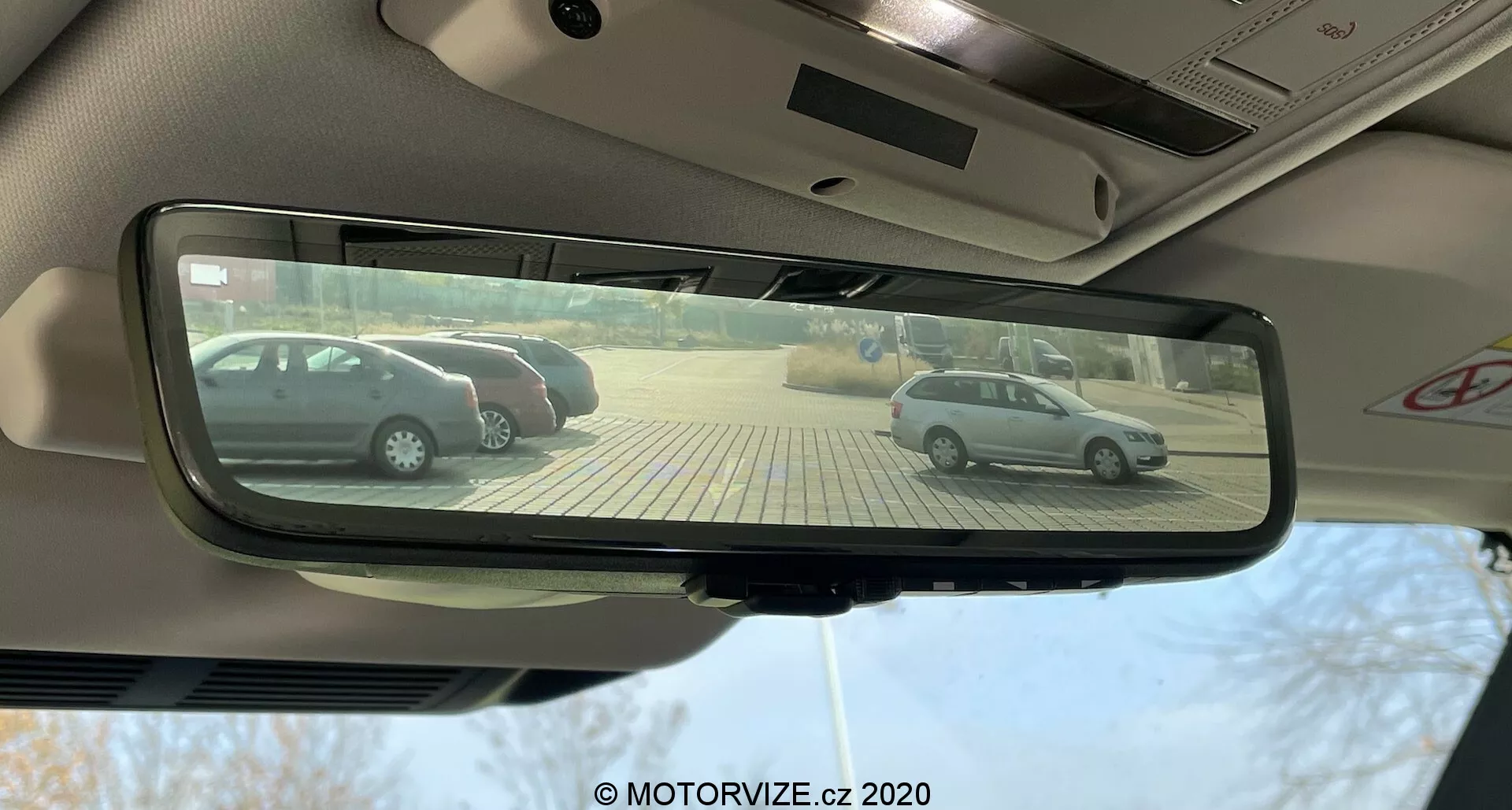 Vista desde el asiento del conductor de un Land Rover Defender 2020, centrada en el espejo retrovisor interior rectangular con atenuación automática, que refleja dos hatchbacks plateados aparcados perpendicularmente en un aparcamiento pavimentado con adoquines beige en un día soleado.