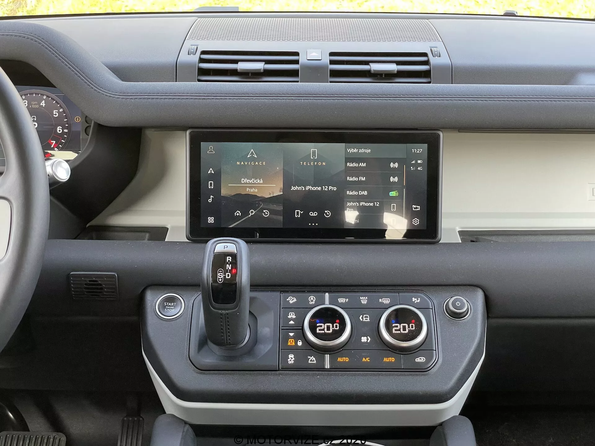Vista del tablero interior del Land Rover Defender 2020 que muestra el sistema de infoentretenimiento con pantalla táctil, salidas de aire dobles, selector de marchas moderno, panel de control de clima de doble zona, vistazo al volante y una combinación de plásticos duros y materiales al tacto suave con un contraste de color en el tablero.
