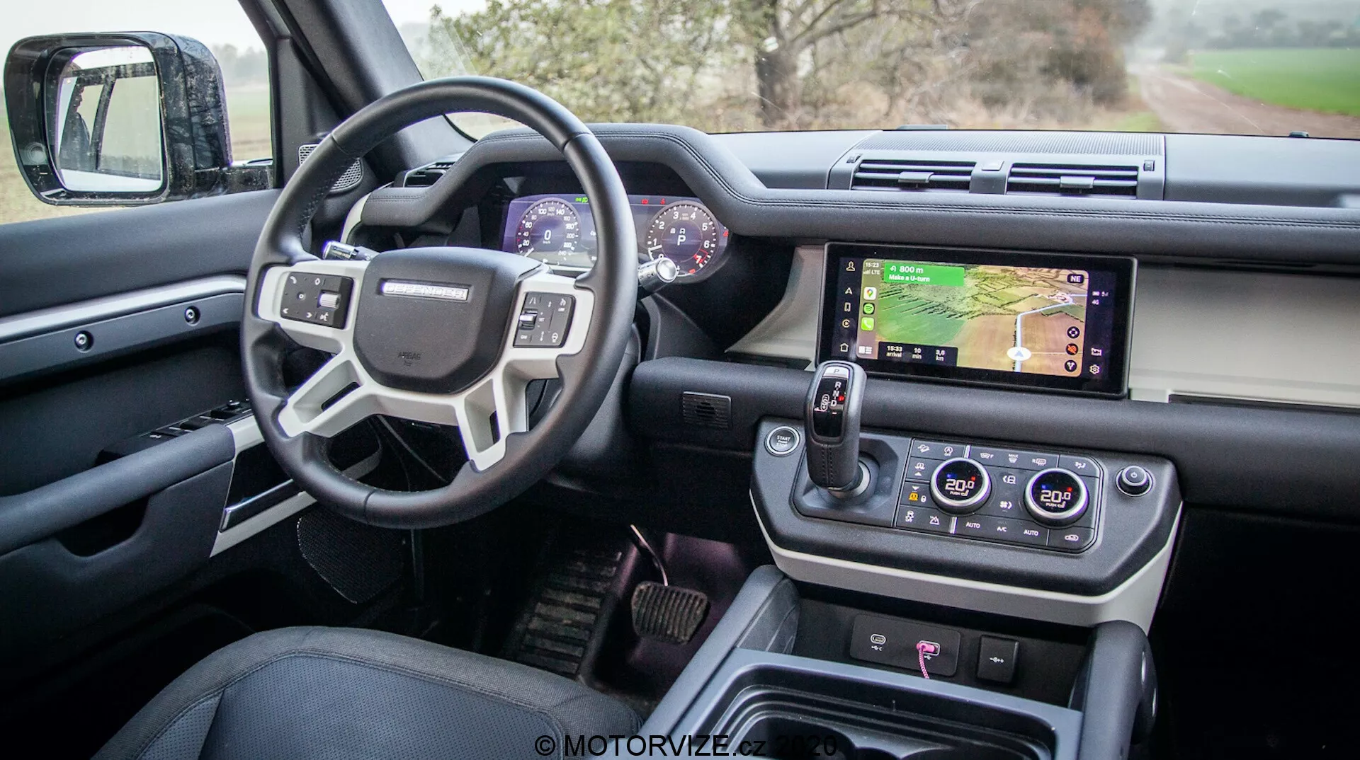 Vista interior de un moderno Land Rover Defender 2020 desde la puerta del conductor mostrando el diseño sofisticado y las características avanzadas, incluyendo una puerta del conductor abierta con controles para ventanas y espejos, un volante multifuncional con botones de control, un cuadro de instrumentos digital, un sistema de infotainment central con pantalla táctil que muestra un mapa, diales para el control del clima, un selector de marchas rotativo moderno y botones de control de terreno, todo en un acabado de lujo y alta calidad.