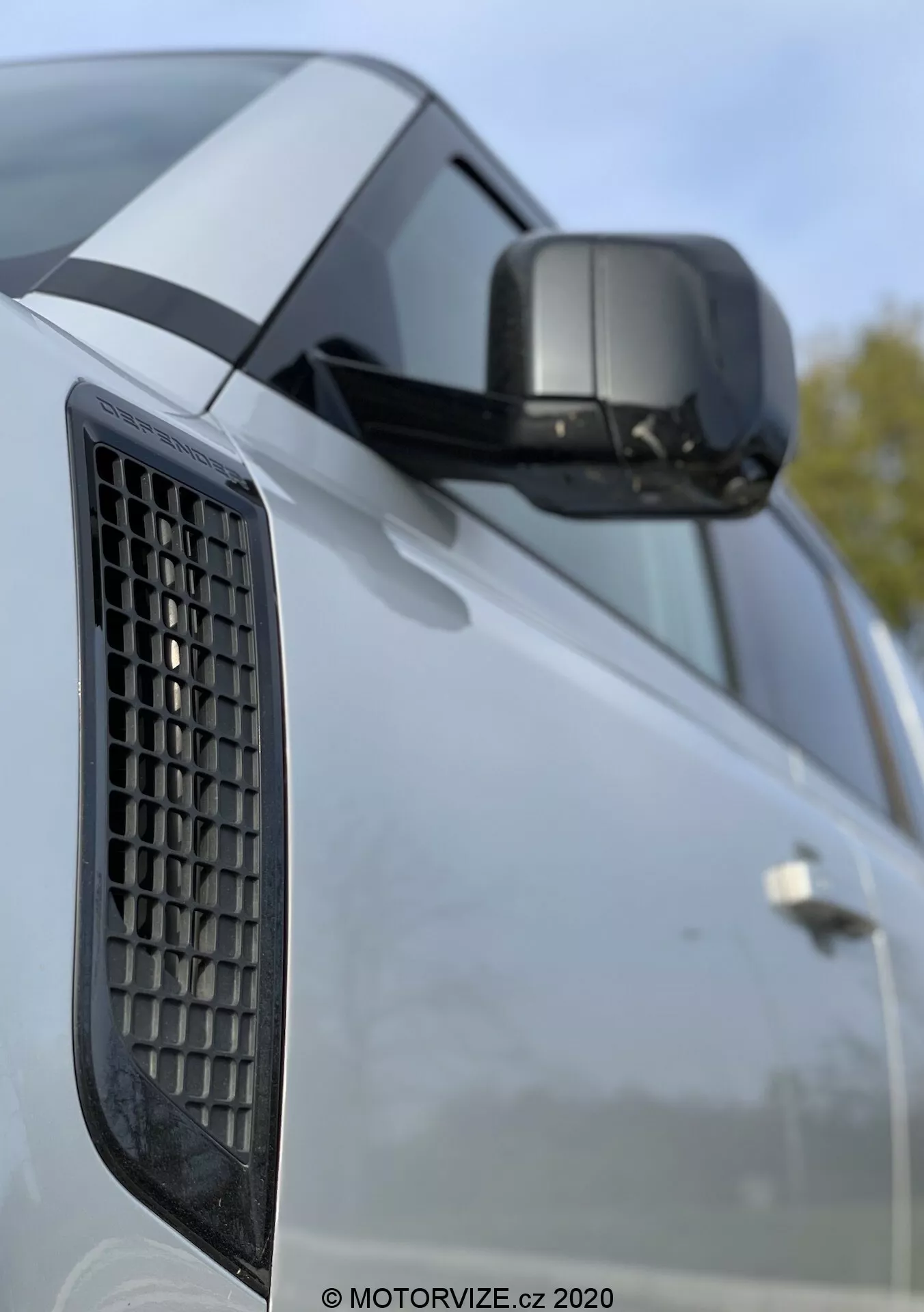 Primer plano del Land Rover Defender 2020 destacando el guardabarros lateral con una rejilla, el espejo lateral con indicador de giro integrado y una parte de la puerta con líneas de carácter pronunciadas; la imagen se centra en los elementos de diseño del vehículo con un fondo desenfocado que sugiere un entorno exterior.