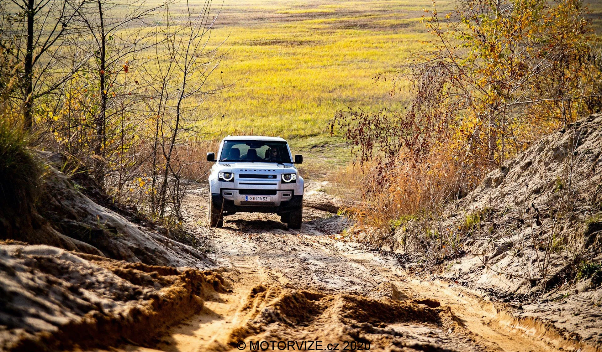 Un SUV Land Rover Defender 2020 se ve visto de frente conduciendo fuera del camino, ubicado centralmente contra un paisaje de terreno desigual y entorno natural. La parte frontal visible del coche muestra la articulación avanzada de la suspensión, los faros LED y una parrilla característica con listones horizontales. Capturando el SUV en un entorno rural con matices otoñales y luz natural, la perspectiva realza la presencia robusta y aventurera del vehículo.