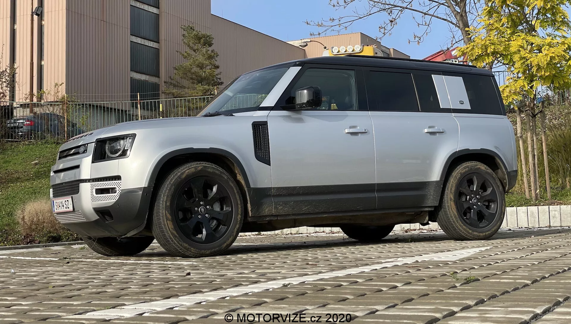 Fotografía de un moderno SUV Land Rover Defender 2020 tomada desde un ángulo bajo para acentuar su postura robusta e imponente. El vehículo se muestra en una vista de tres cuartos desde el frente y el costado, destacando su parrilla distintiva, faros LED, parachoques con luces antiniebla y sensores, llantas de aleación negras con diseño multirradio y neumáticos de perfil bajo, espejos laterales negros con tecnologías adicionales potenciales, ventanas tintadas, línea de techo prominente, manijas de puertas al ras, fuerte diseño de arcos de rueda y color plateado o gris claro contrastado con elementos de adorno negro. El SUV está estacionado sobre un suelo adoquinado con un edificio y follaje en el fondo bajo iluminación diurna.