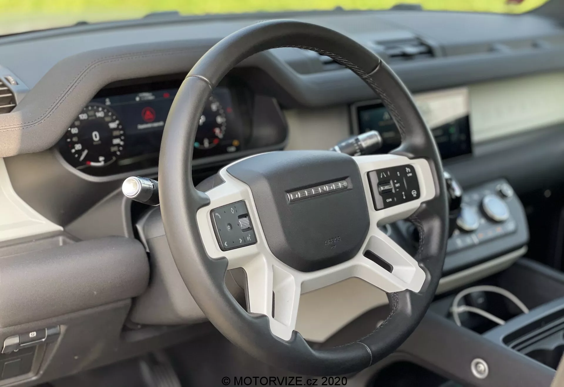 Vista interior del Land Rover Defender 2020 centrada en el volante y el tablero desde la perspectiva del asiento del pasajero delantero. El volante moderno de tres radios cuenta con controles multifunción y un panel de instrumentos digital para la visualización clara de los indicadores. La combinación de materiales como cuero y adornos metálicos o plásticos destaca el diseño ergonómico y tecnológico del vehículo.