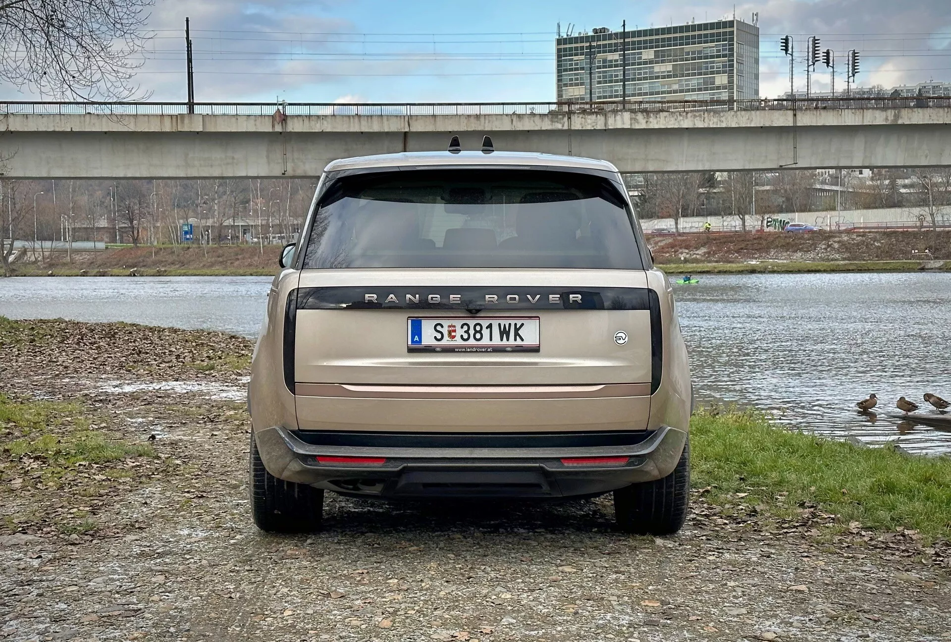 Vista trasera de Range Rover 2021 con ángulo de visión directo a nivel de ojos, aparcado cerca de un cuerpo de agua con patos y un paso elevado al fondo, mostrando el nombre del modelo, luces traseras, sensores de aparcamiento y sistema de escape doble en beige mate