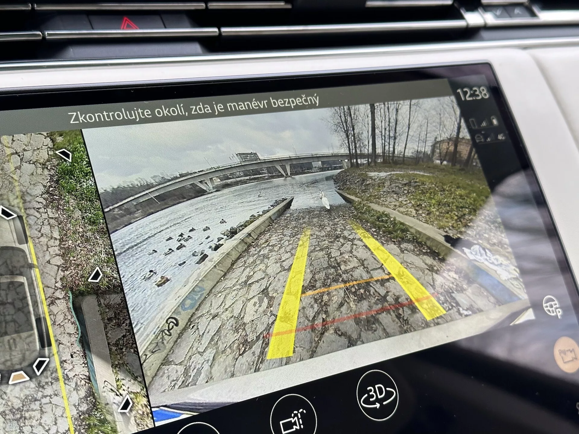 Pantalla de infotenimiento de Land Rover Range Rover 2021 mostrando vista trasera desde cámara de reversa con líneas guías dinámicas en un área de estacionamiento cerca de una zona acuática, detallada con vegetación, íconos de asistencia de estacionamiento y texto de recordatorio de seguridad