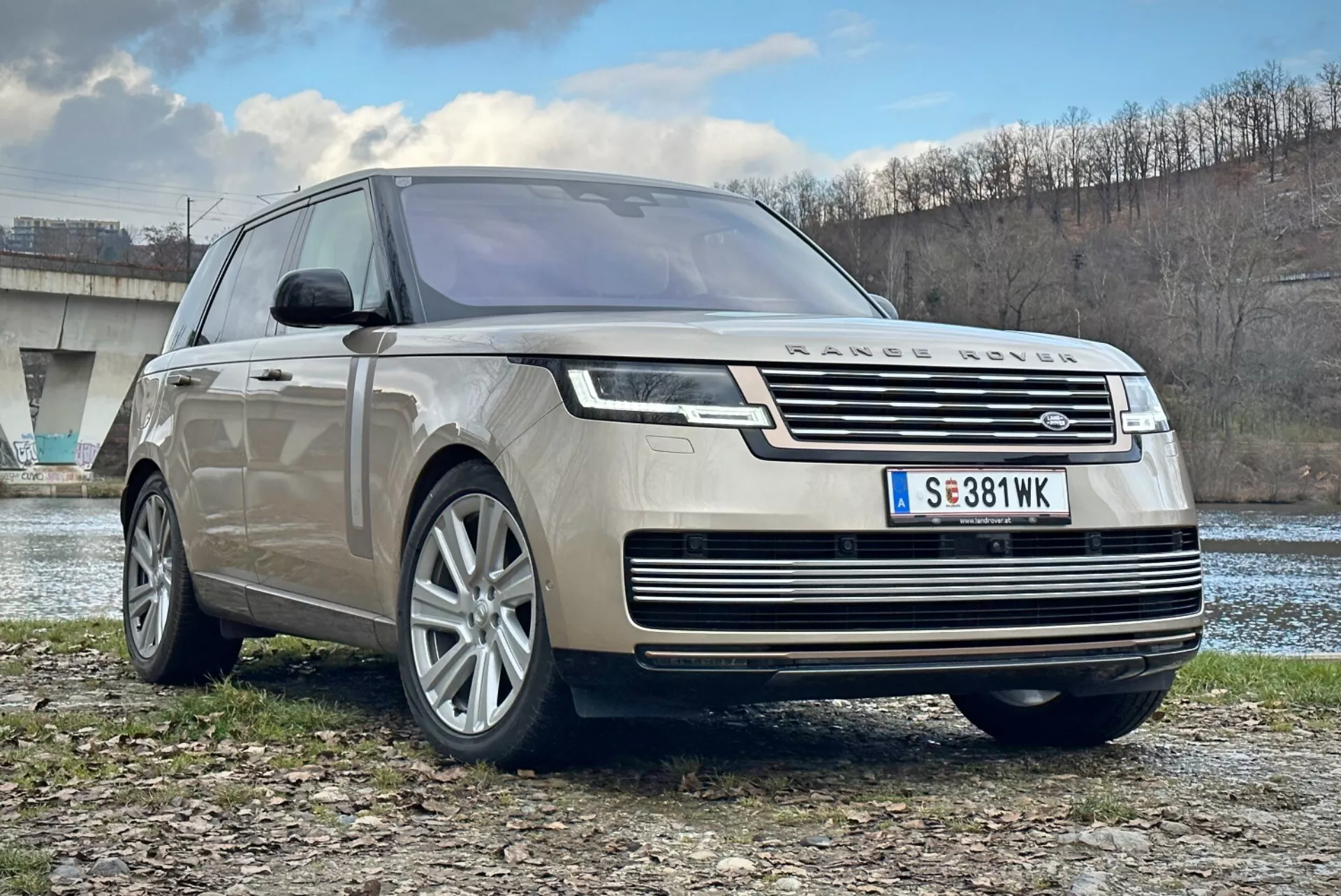 SUV de lujo Land Rover Range Rover 2021, vista frontal de tres cuartos destacando la parrilla cromada, faros LED, ruedas de aleación de múltiples radios y diseño aerodinámico, estacionado al aire libre cerca de un río y un puente