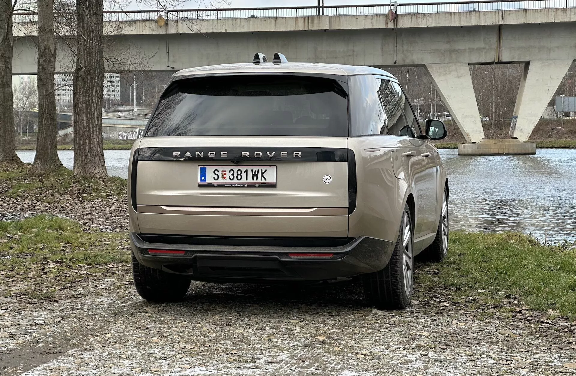 Vista trasera de un Range Rover 2021 aparcado al aire libre con un cuerpo de agua y un puente al fondo, mostrando luces traseras LED, escape doble, cristales tintados y llantas de aleación de múltiples radios.
