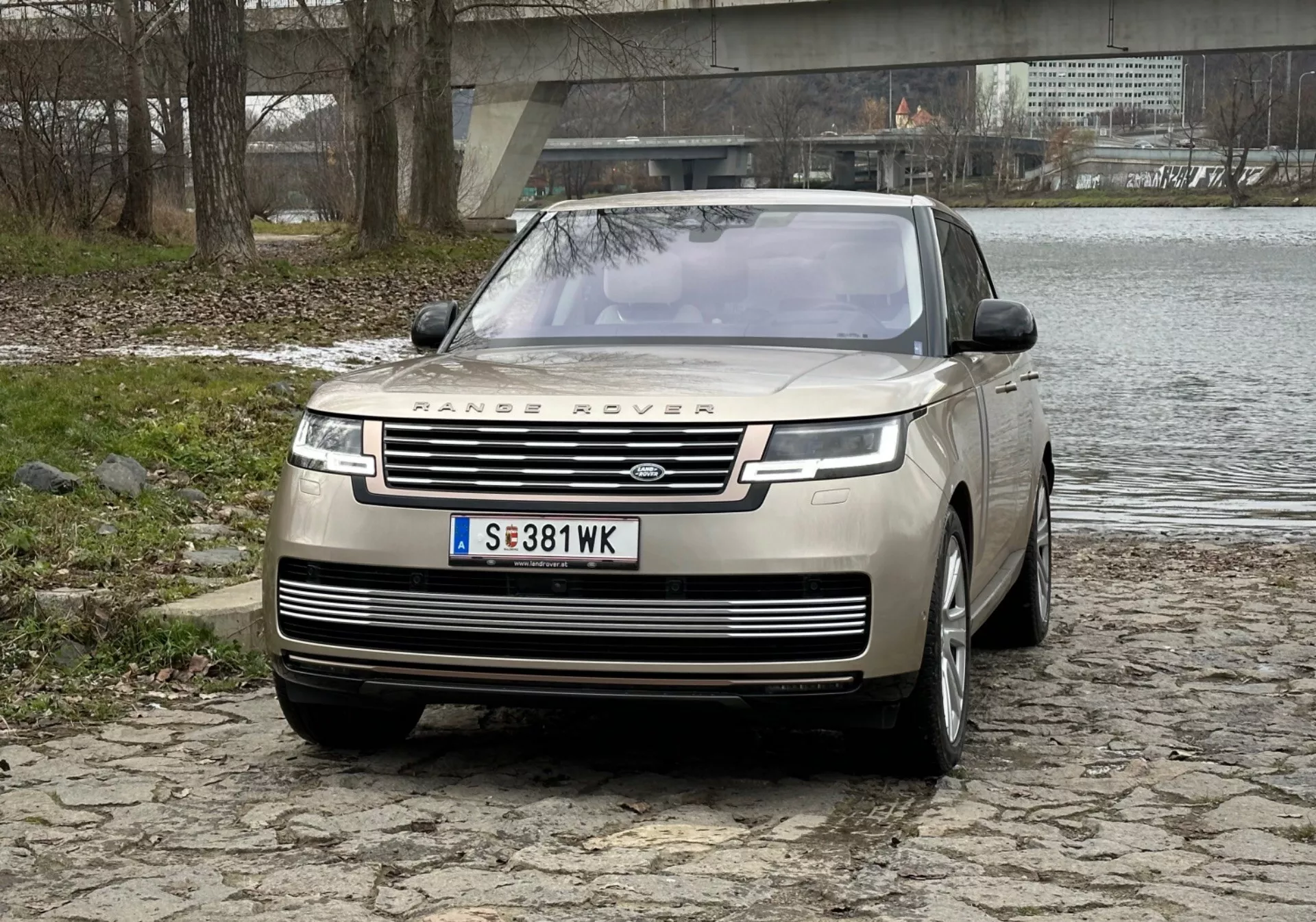 SUV de lujo Range Rover 2021 en plata, mostrando parrilla frontal prominente con marca, luces diurnas LED elegantes, paragolpes delantero con sensores de estacionamiento y diseño de capó clamshell en superficie adoquinada cerca de un río