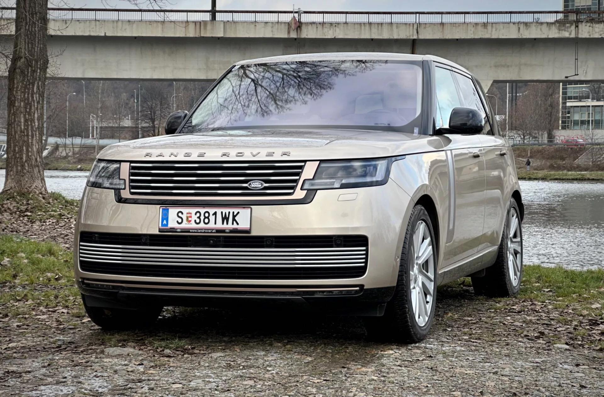 Range Rover 2021 en una impresionante vista frontal de tres cuartos que muestra su distintiva parrilla, faros delanteros LED elegantes y robusta placa protectora frontal contra un fondo de parque urbano, enfatizando el lujo y la capacidad todoterreno.