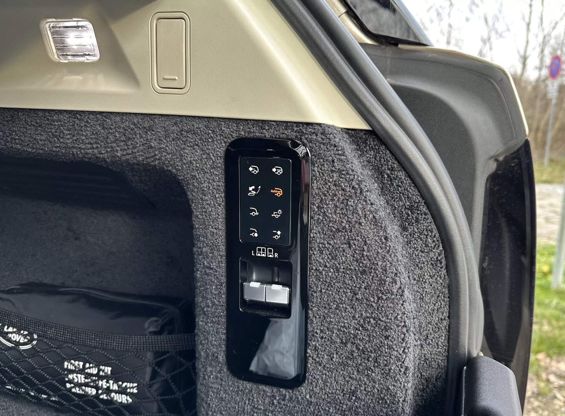 Vista detallada del panel de control electrónico del maletero en el interior de un Land Rover Range Rover 2021, mostrando botones para operaciones como la apertura y cierre electrónicos, control de la cubierta de carga, posiblemente una luz de maletero o toma de corriente, ajustes de los asientos traseros desde el maletero y liberación de emergencia, situado contra la pared lateral alfombrada del maletero y el cielo raso de color claro con luminaria de carga.