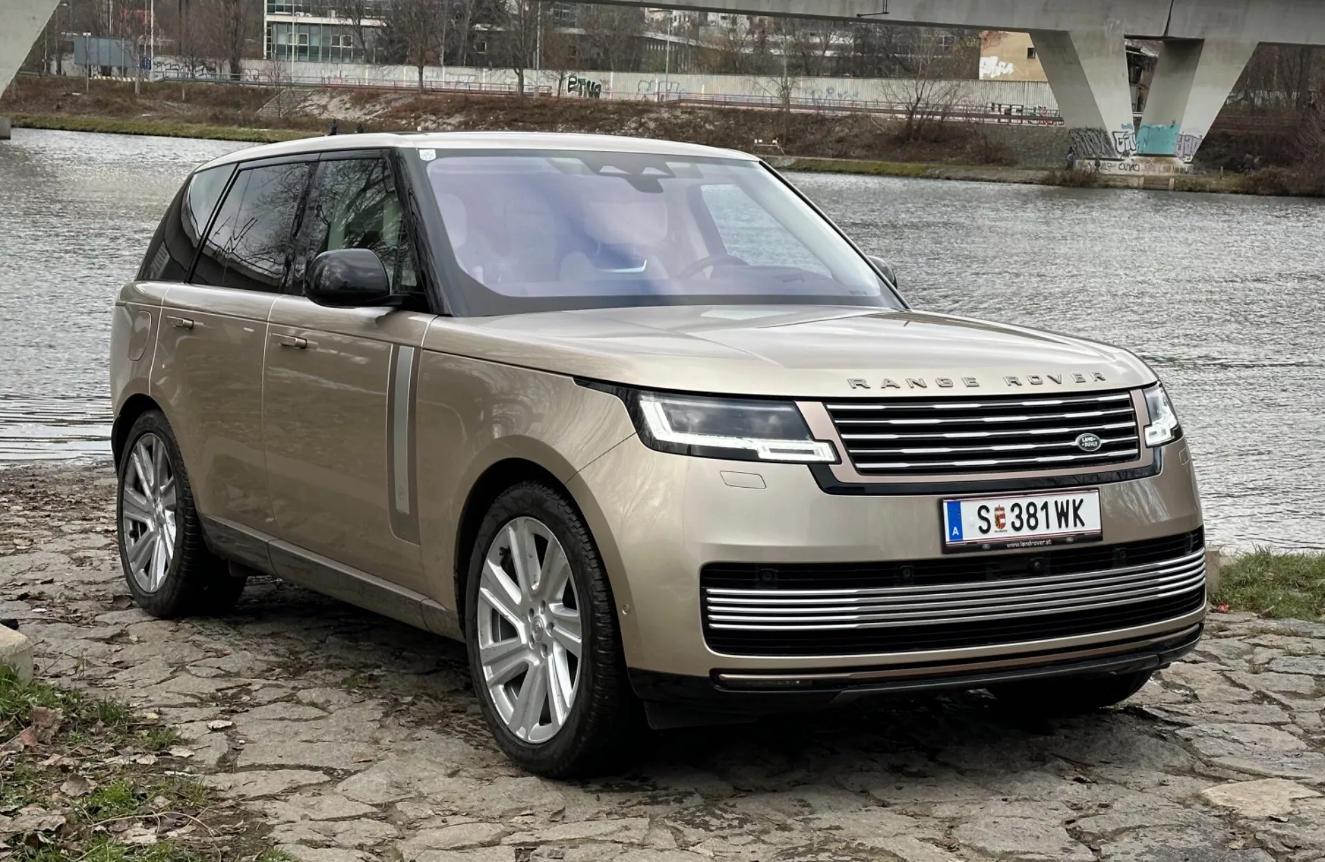 SUV de lujo Land Rover Range Rover 2021 estacionado junto a un río con un puente al fondo, mostrando su diseño moderno y características tecnológicas incluyendo faros LED, parrilla frontal con emblema, llantas de aleación de múltiples radios y techo solar panorámico.
