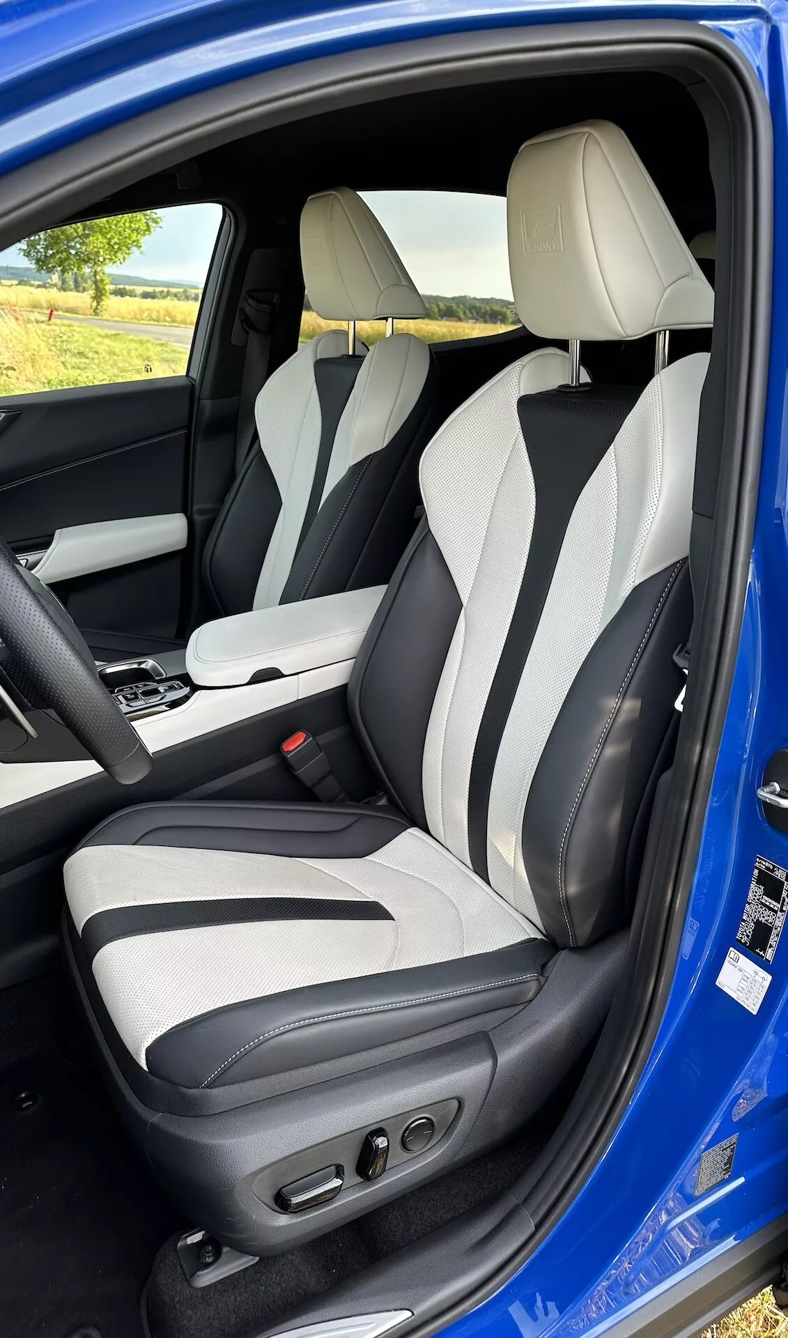 Vista interior a través de la puerta abierta del lado del conductor de un Lexus NX 2021, mostrando el área del conductor desde una ligera elevación, con tapicería de cuero bicolor, controles avanzados del asiento del conductor, refuerzos laterales, reposacabezas integrados y consola central con palanca de cambios automática