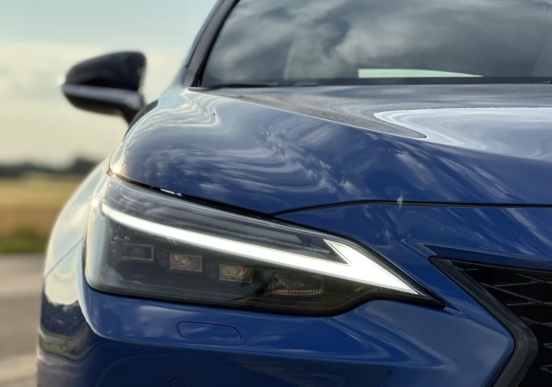 Primer plano del cuarto frontal izquierdo del Lexus NX 2021 con ensamblaje de faro LED, capó esculpido, parachoques delantero con parrilla y acabado azul profundo brillante, enfatizando la tecnología de iluminación avanzada y el diseño aerodinámico.