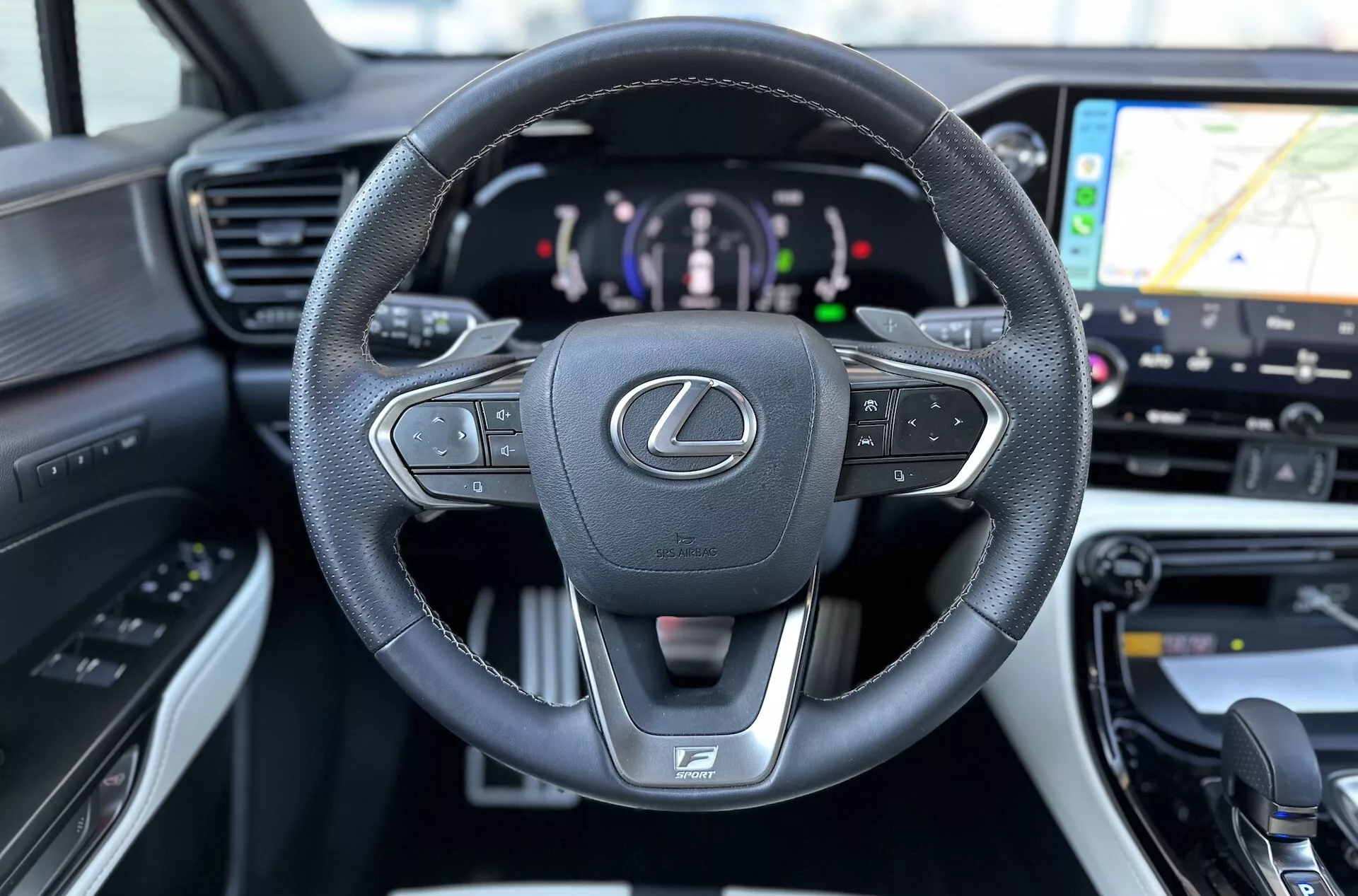 Interior del Lexus NX 2021 enfocado en el volante multifuncional forrado en cuero con emblema Lexus y logo F SPORT, panel de instrumentos digital indicando tren motriz híbrido, y vislumbre del sistema de navegación GPS en el sistema de infotenimiento