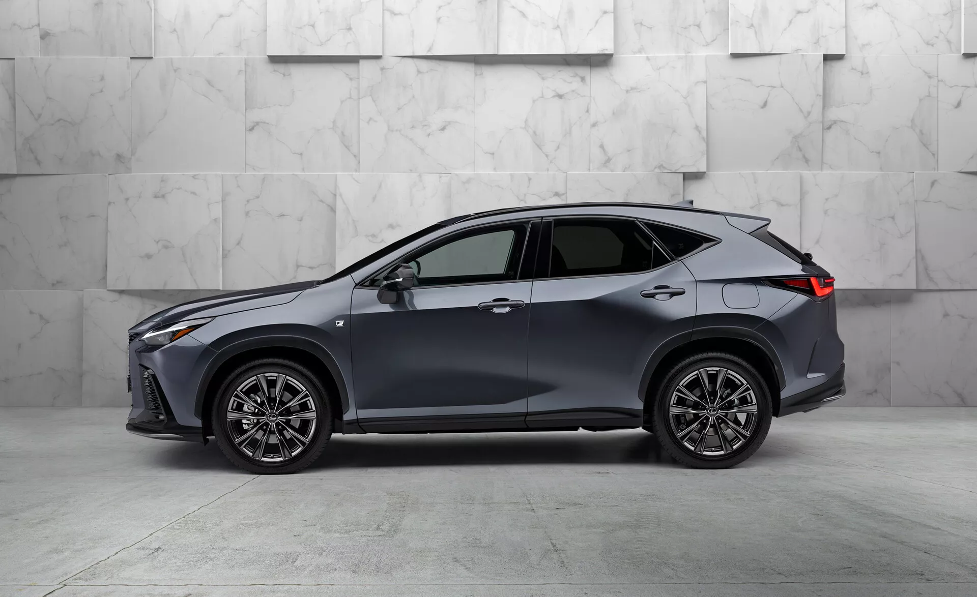 Vista de perfil lateral de un moderno Lexus NX 2021 crossover SUV, que muestra su elevada altura libre al suelo y forma de carrocería. Pintado en un tono oscuro, posiblemente gris o azul, el vehículo presenta cinco puertas, diseño aerodinámico elegante, grandes ruedas de aleación multirradio, delgados faros y luces traseras LED, espejos laterales del mismo color de la carrocería con señales de giro integradas, ventanas con molduras cromadas y una antena de aleta de tiburón, todo ello contra un fondo de mármol.