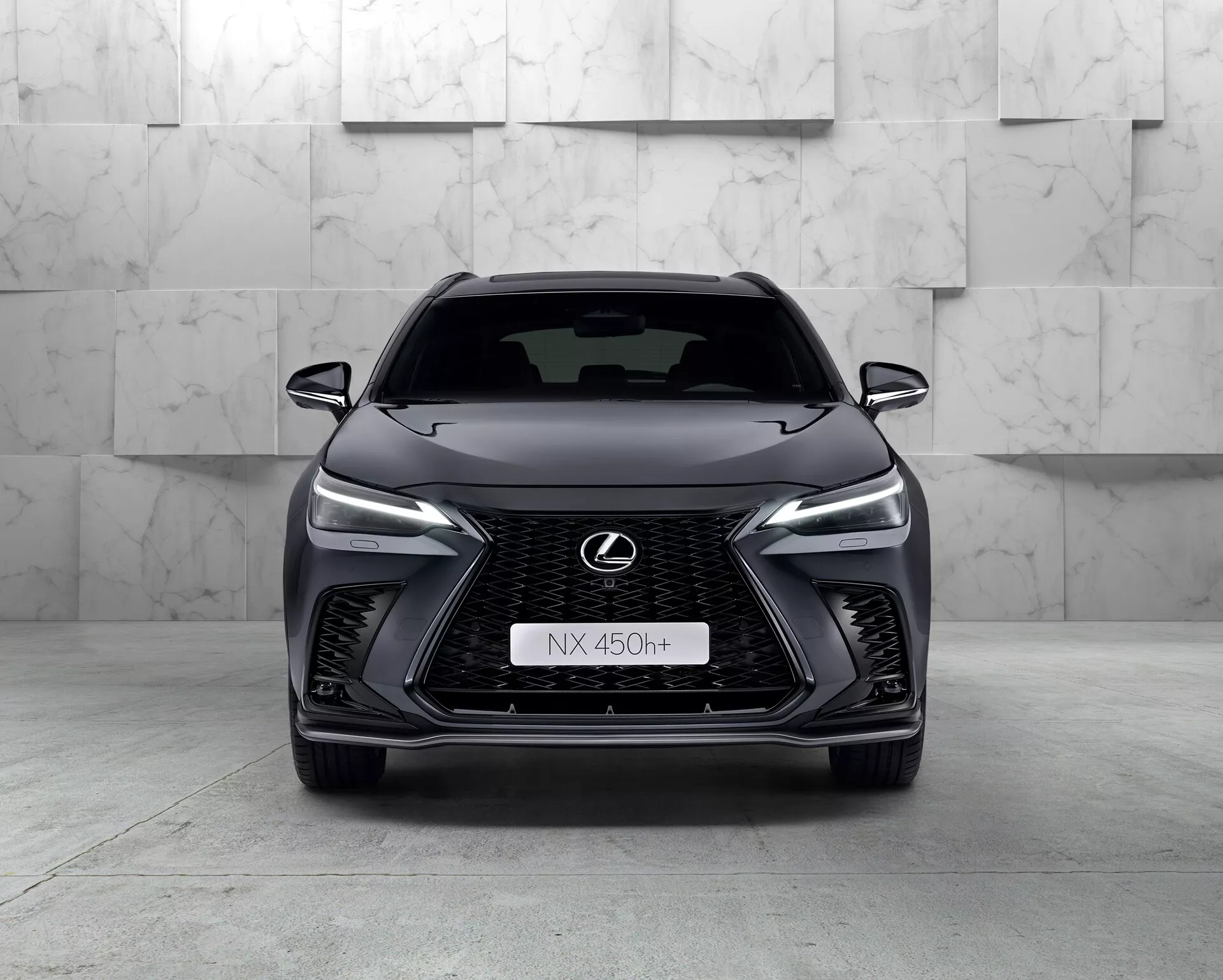 Vista frontal del Lexus NX 450h+ 2021, un SUV moderno y elegante que muestra su prominente parrilla en forma de huso con diseño de malla, faros LED estrechos, emblema de Lexus cromado con contorno azul y diseño agresivo del parachoques delantero. La naturaleza híbrida del coche está indicada por el distintivo 'h+' en la parrilla. El entorno del estudio resalta las curvas y características del vehículo con una iluminación limpia y controlada.