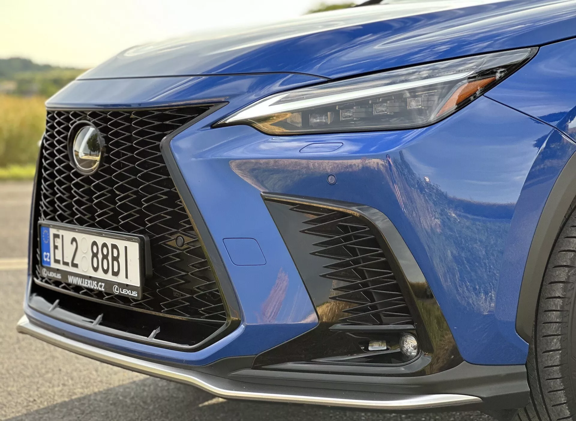 Primer plano de la esquina frontal del Lexus NX 2021 destacando el faro con tecnología LED, diseño distintivo de la rejilla de malla y parachoques delantero aerodinámico con entradas de aire, resaltado por una pintura metálica azul profundo y un vistazo a una rueda de aleación de lujo.