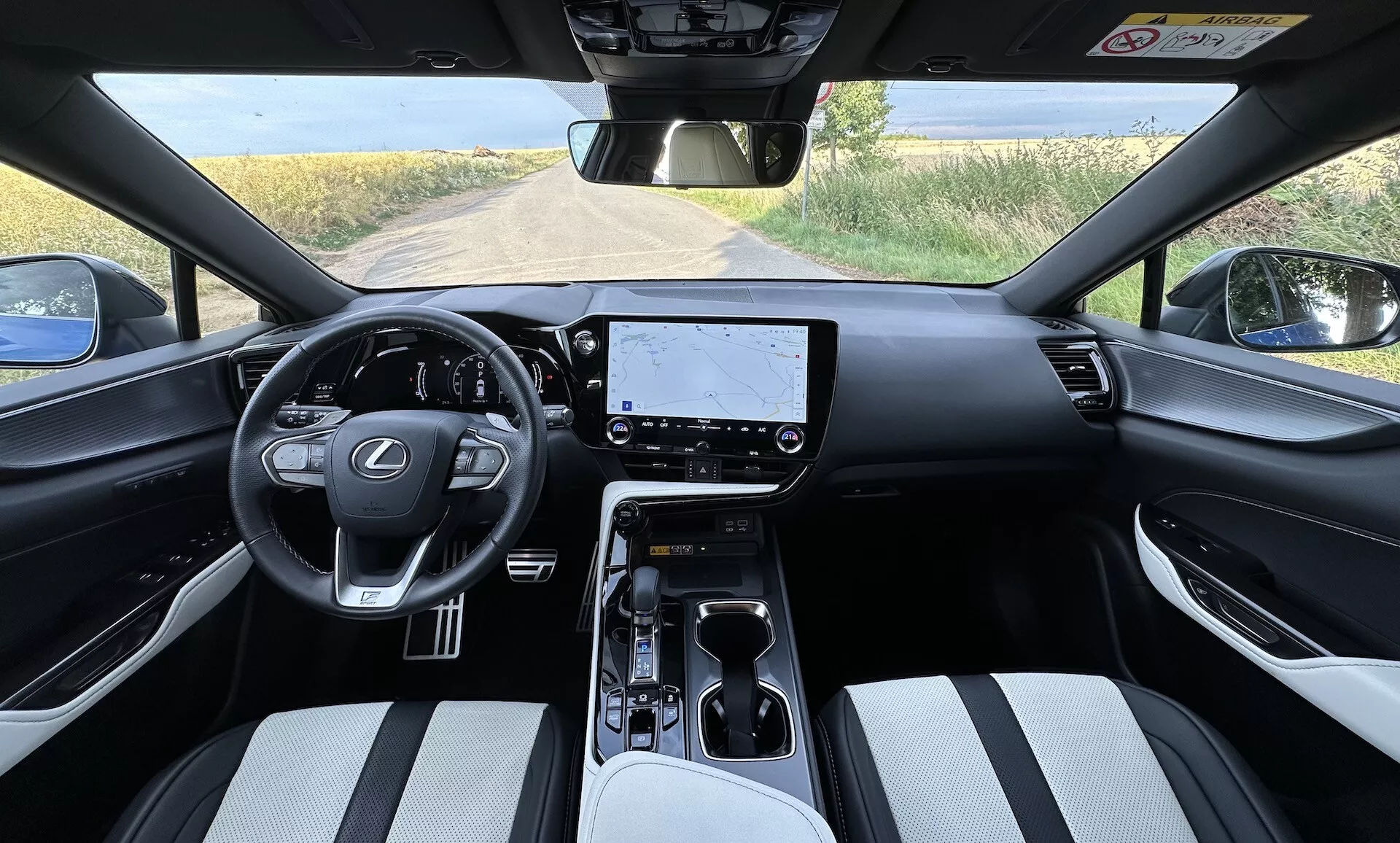 Vista interior del Lexus NX 2021 desde la perspectiva del conductor con volante centrado con logo de Lexus y botones de control, una gran pantalla táctil de infotainment en el tablero, controles climáticos minimalistas, selector de cambio automático en la consola central con portavasos y almacenamiento, asientos tapizados en material bitono, y una vista parcial de un paisaje rural a través del parabrisas.