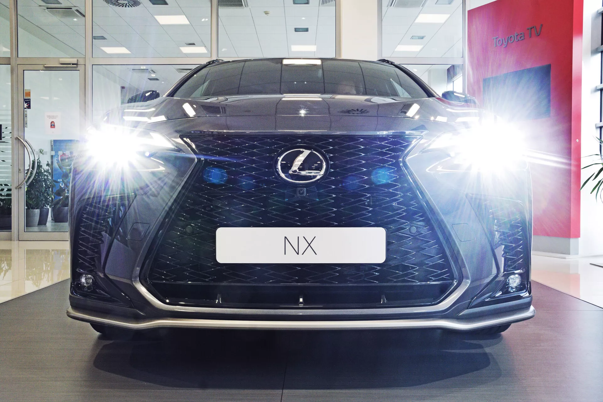 Vista frontal del vehículo Lexus NX 2021 fotografiado desde un ángulo bajo, destacando su altura y presencia en la carretera en un entorno de sala de exposición. La imagen resalta la distintiva parrilla de husillo con forma de reloj de arena y malla oscura, los faros LED angulares iluminados con efectos de resplandor, luces diurnas y detalles del parachoques inferior. El suelo pulido y el soporte de placa de matrícula con el letrero 'NX' añaden al atractivo lujoso y tecnológico del coche.
