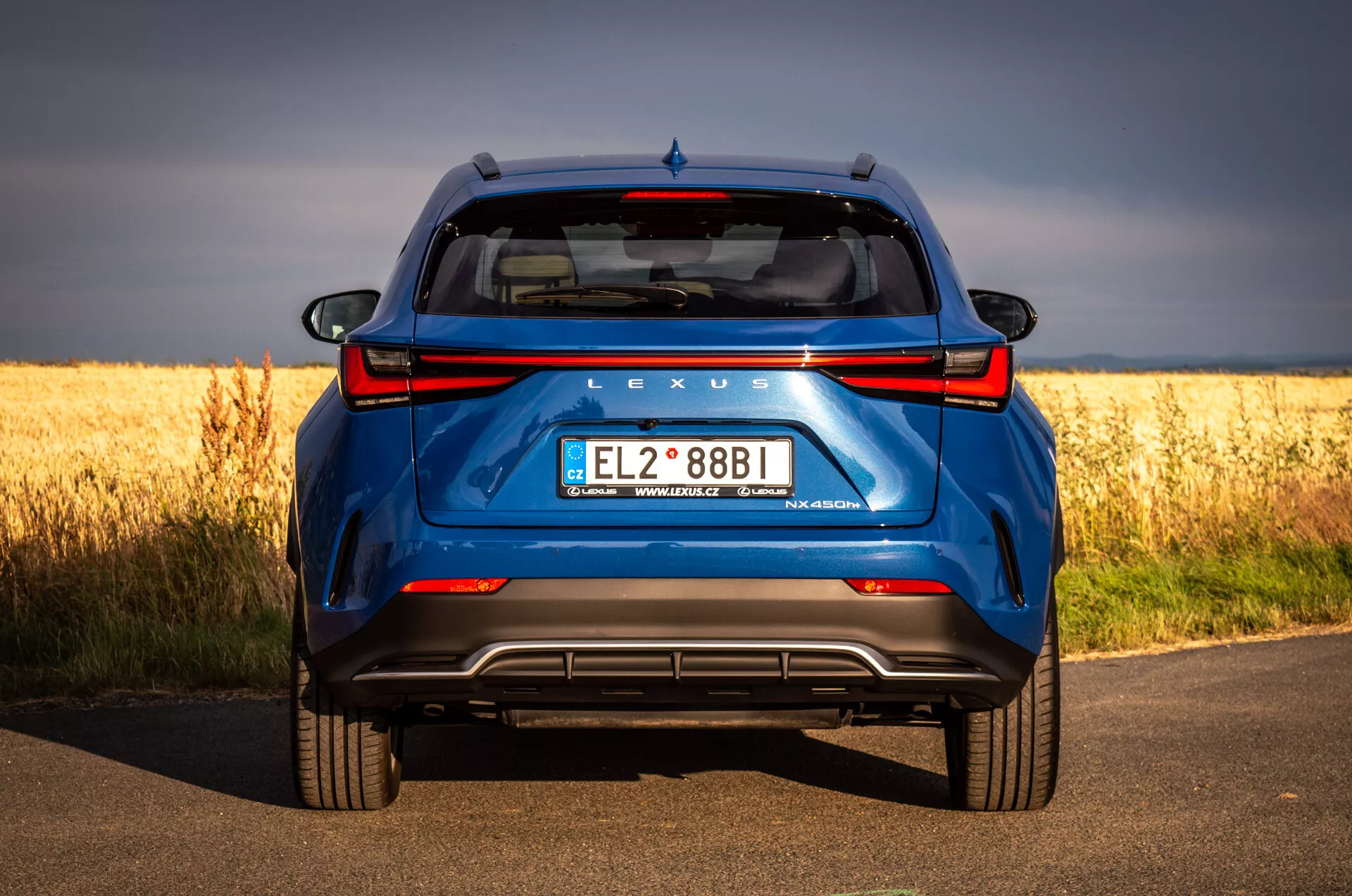 Vista trasera de SUV azul Lexus NX 2021 en carretera de asfalto con campo dorado en el fondo, destacando la tecnología híbrida y las características de diseño