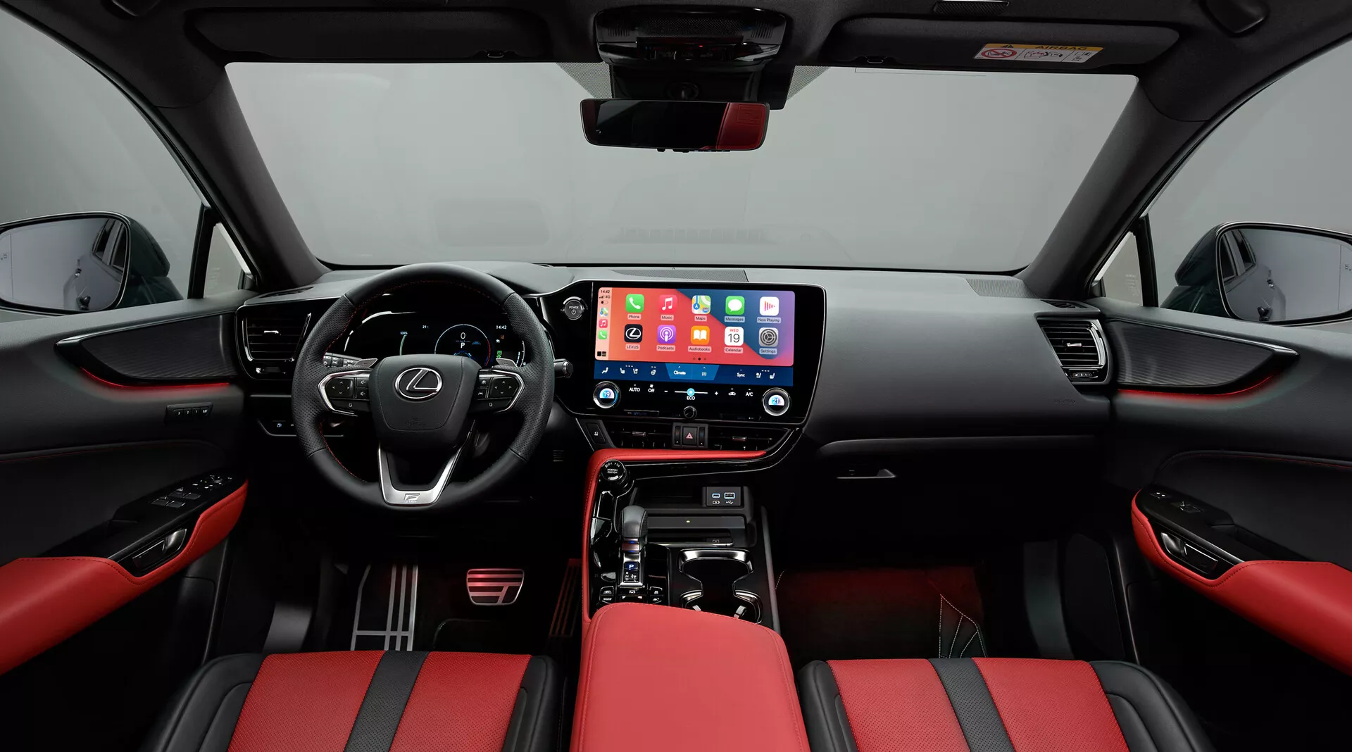 Interior del Lexus NX 2021 enfocado en la cabina delantera, tomado desde una posición central. La perspectiva de ángulo amplio muestra el tablero con una gran pantalla táctil central de infoentretenimiento que muestra una interfaz móvil, controles físicos del clima y una palanca de cambios para la transmisión automática. El volante multifuncional con botones de control sugiere características avanzadas. Los asientos están tapizados en cuero bicolor rojo y negro, proporcionando un aspecto deportivo. Los paneles de las puertas combinan con el tema rojo y negro con controles de ventanas eléctricas y una rejilla de altavoz visible, enfatizando la modernidad y el lujo.