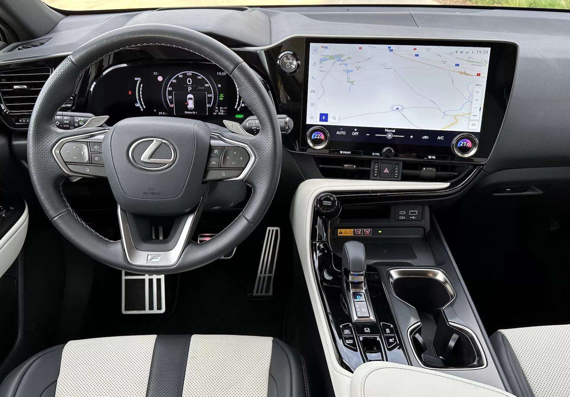 Vista interior del Lexus NX 2021 mostrando el área del conductor y la consola central desde un ángulo elevado, con el logotipo de Lexus en el volante con controles montados, un cuadro de instrumentos digital iluminado, un gran sistema de infotenimiento táctil con navegación GPS, botones de control de clima y audio, selector de marchas ergonómico, controles de modo de conducción, materiales de lujo en asientos y puntos de contacto, acentos metálicos, y portavasos visibles y compartimento de almacenamiento.