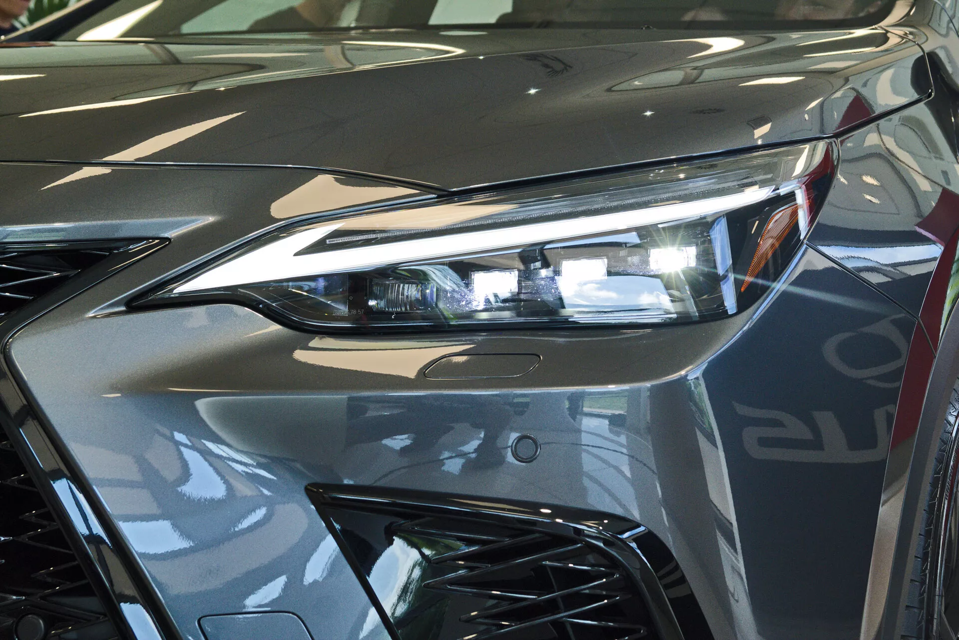 Vista de cerca del extremo delantero de un Lexus NX 2021, enfocándose en el ensamblaje del faro del lado del conductor con tecnología avanzada de iluminación LED y parte de la parrilla de estilo agresivo. La imagen capta el brillo reflectante de la pintura plateada metálica y las líneas dinámicas que contribuyen a la estética aerodinámica del vehículo. Fotografiado en interiores, probablemente en una sala de exposición, enfatizando el diseño lujoso y el acabado de alta calidad del vehículo.