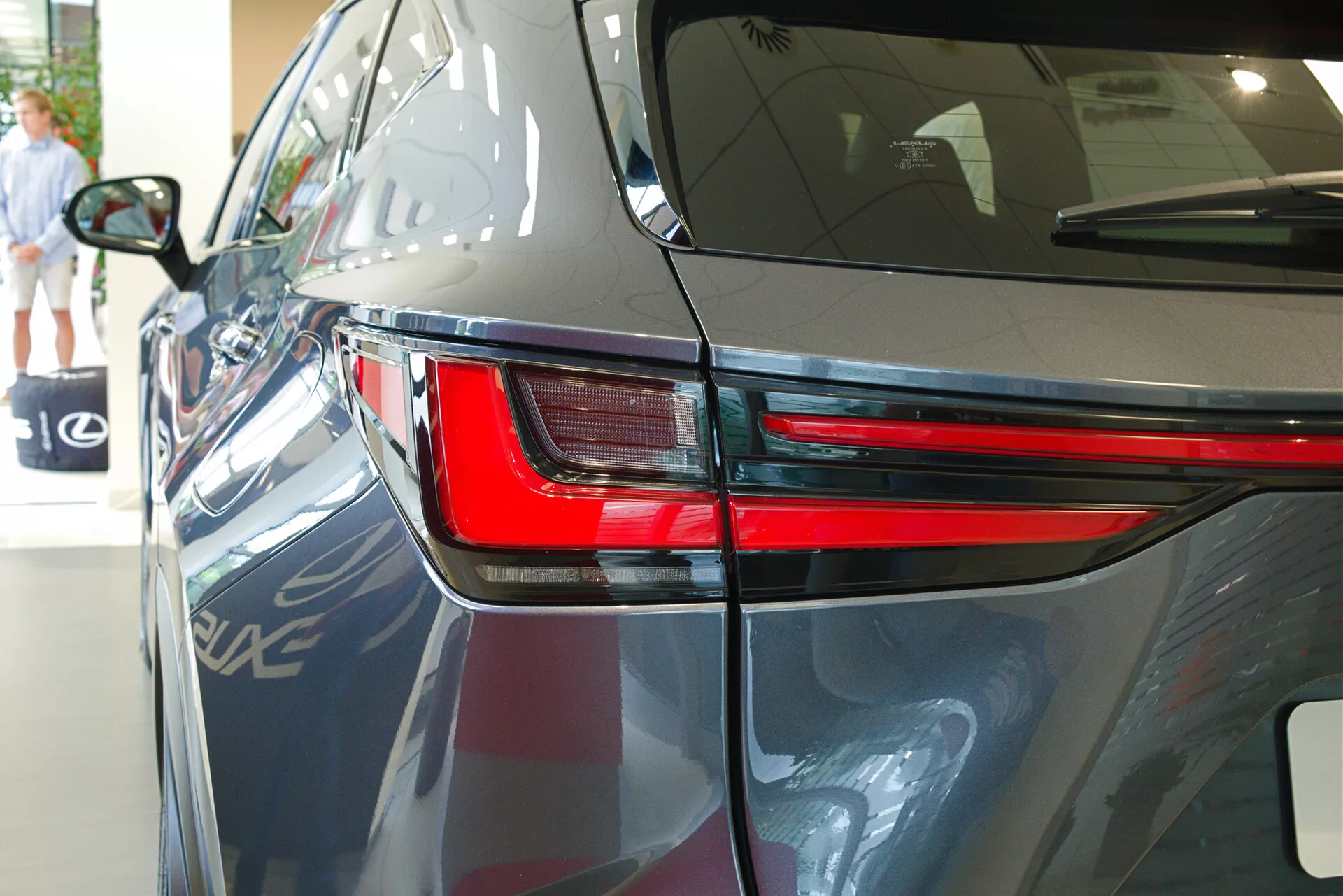 Primer plano del Lexus NX 2021 desde un ángulo trasero de tres cuartos, destacando el elegante diseño de la luz trasera LED, parte del panel trasero y una porción de la tapa del maletero. La imagen muestra la pintura oscura y brillante del automóvil, las líneas aerodinámicas del cuerpo y sugiere un entorno de exhibición interior con reflejos de otro vehículo, elementos arquitectónicos y una persona caminando en el fondo.