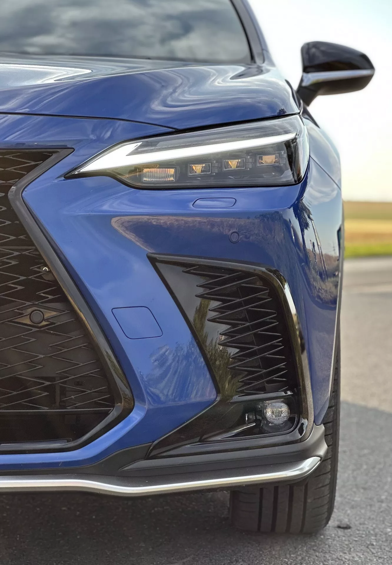 Primer plano de la esquina delantera izquierda de un Lexus NX de 2021 en azul oscuro destacando el avanzado diseño de faros LED, intrincado patrón de parrilla, paragolpes delantero deportivo y posibles características adaptativas de iluminación y aerodinámicas con un acabado metálico brillante