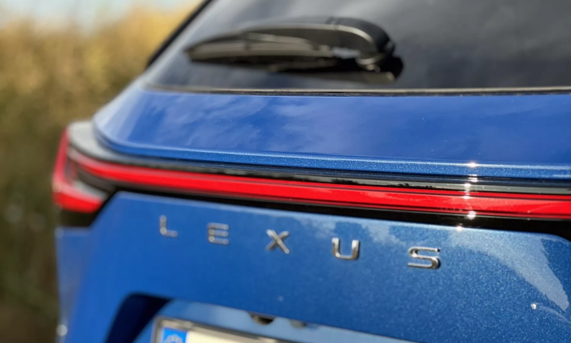 Vista de cerca de la zona de marca trasera del Lexus NX 2021 con distintivo Lexus plateado, una tira de luz roja sobre el nombre, en un cuerpo azul metálico bajo condiciones de luz diurna